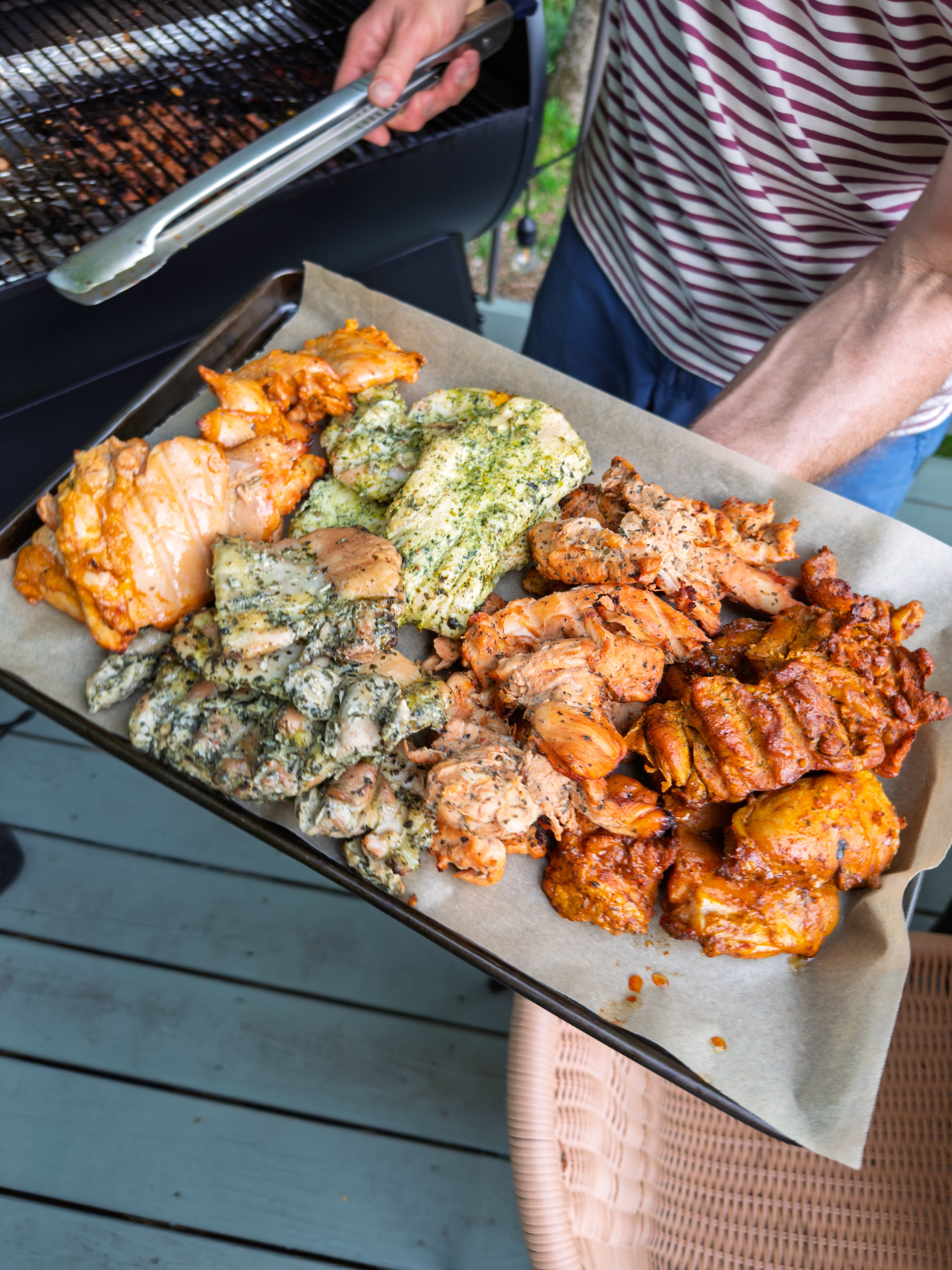 Person holding a tray of grilled meats with various seasonings, standing near a grill on a wooden deck