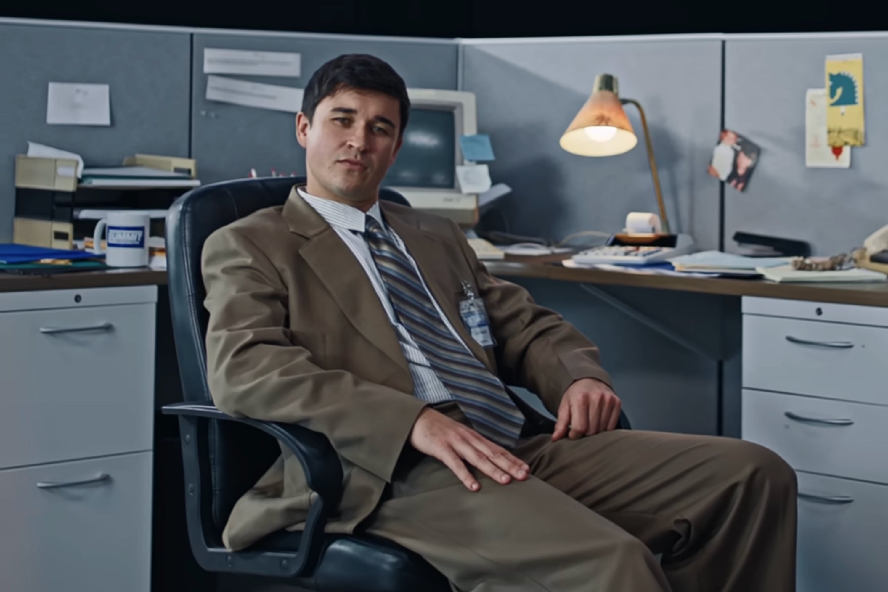 Person in a business suit sits casually in an office cubicle, with a relaxed posture and papers scattered on the desk