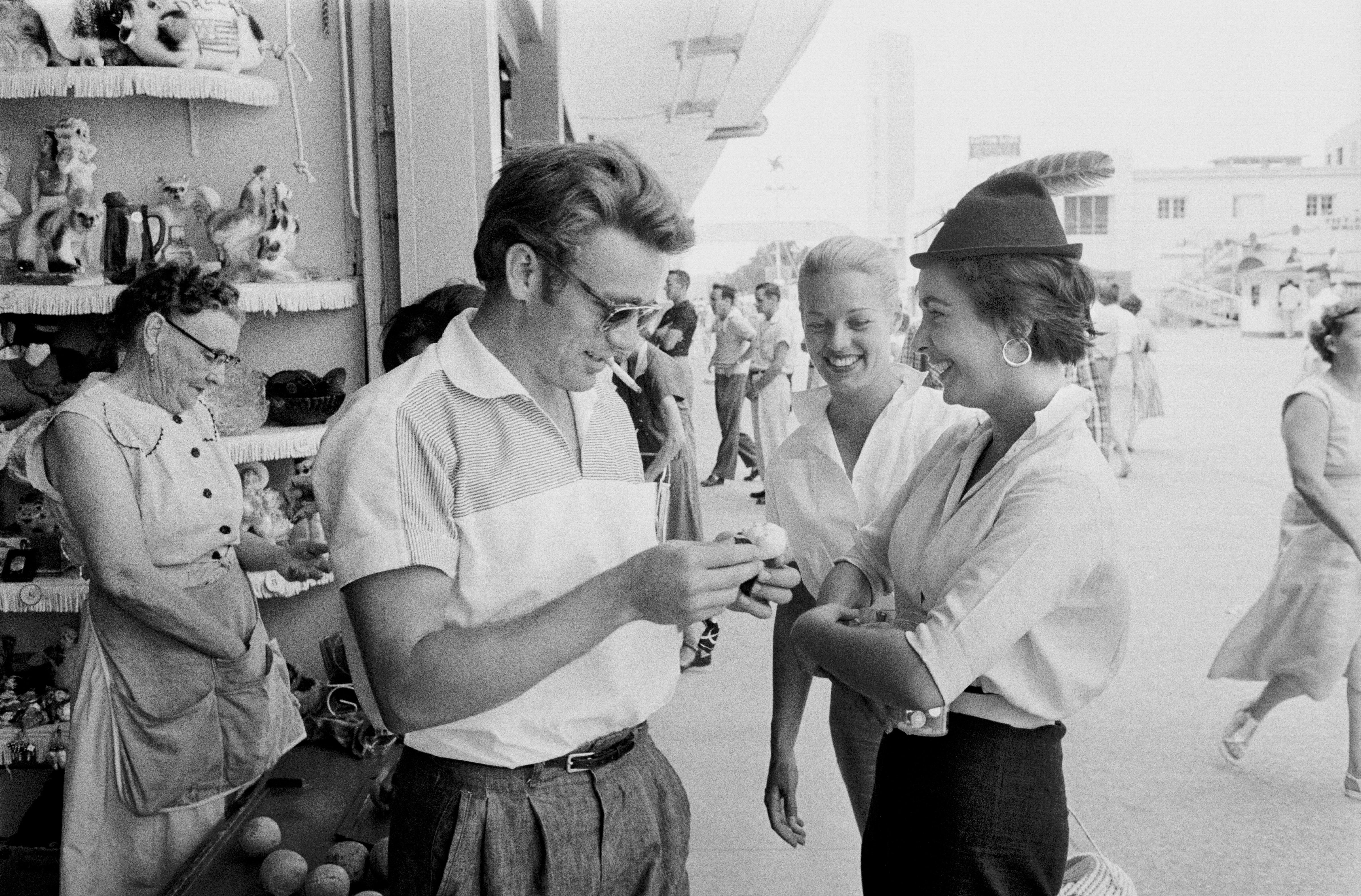 James Dean, Pat Westmore and Elizabeth Taylor smiling in casual attire stand near a shop with displayed items, engaged in a light conversation