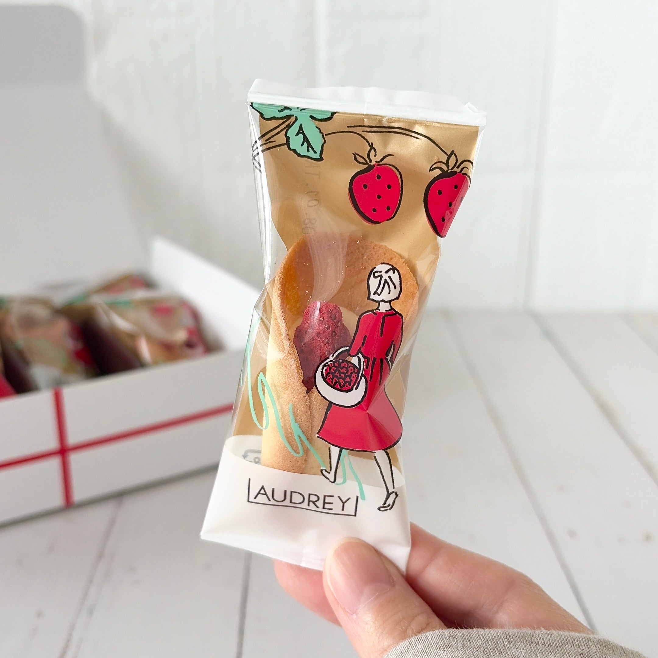 オードリー「グレイシアミルク」イラスト入りパッケージのストロベリー菓子を手に持っている様子。背景には同様の箱に入った商品が見える。
