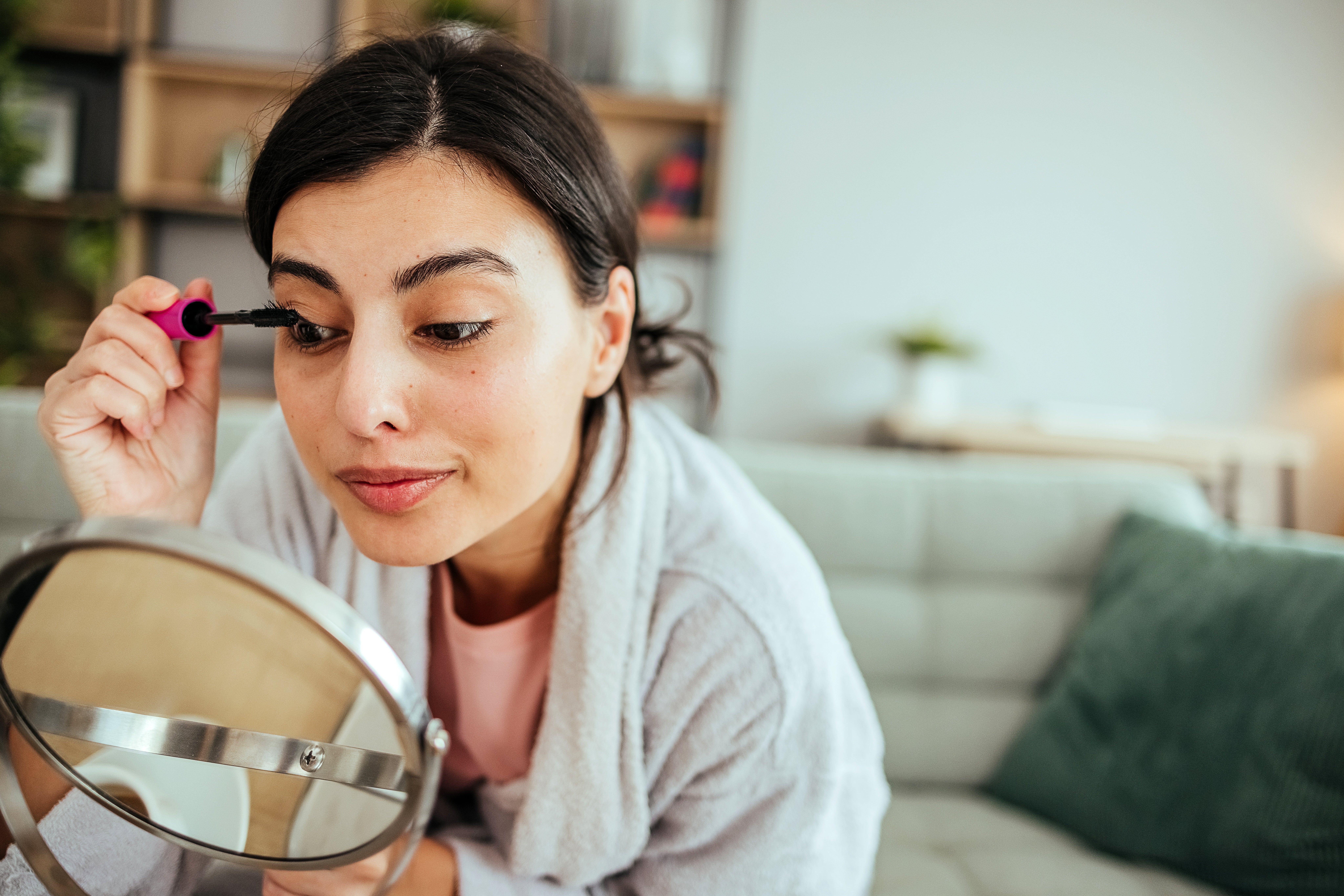 A woman applying mascara in front of a round mirror, wearing a cozy robe, seated on a couch in a living room setting with shelves in the background