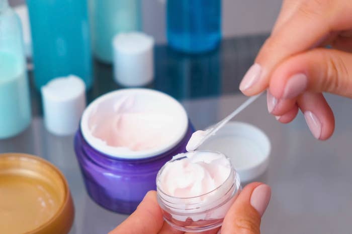A person scooping cream from a small container using a spatula, surrounded by various skincare jars