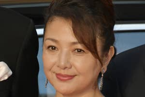 女性がレッドカーペットでシンプルなデザインのドレスを着て微笑んでいる。髪はアップスタイルで、長いピアスをつけている。