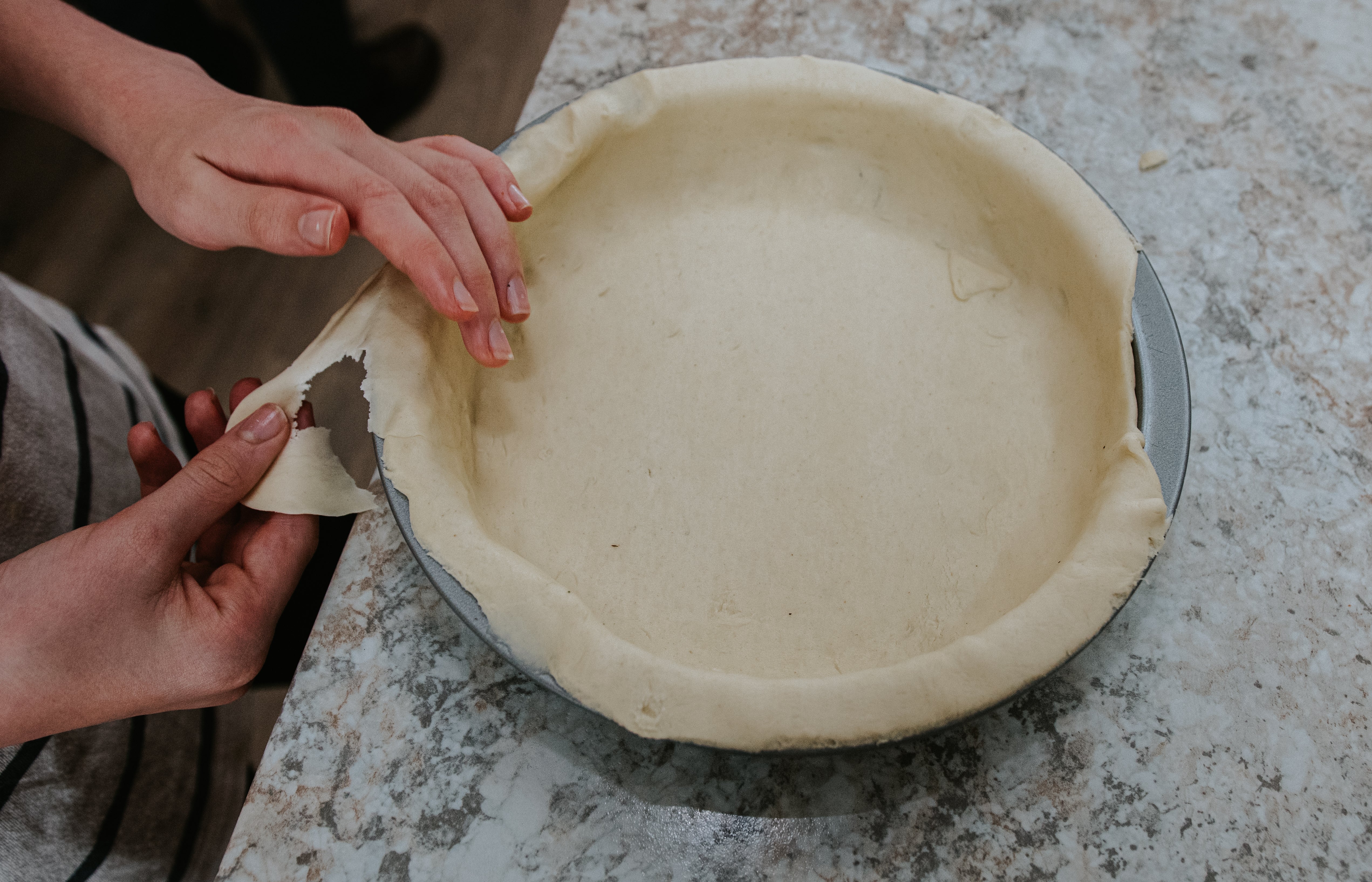 Person preparing a pie crust in a baking dish on a countertop. Hands are shaping and trimming the dough edges