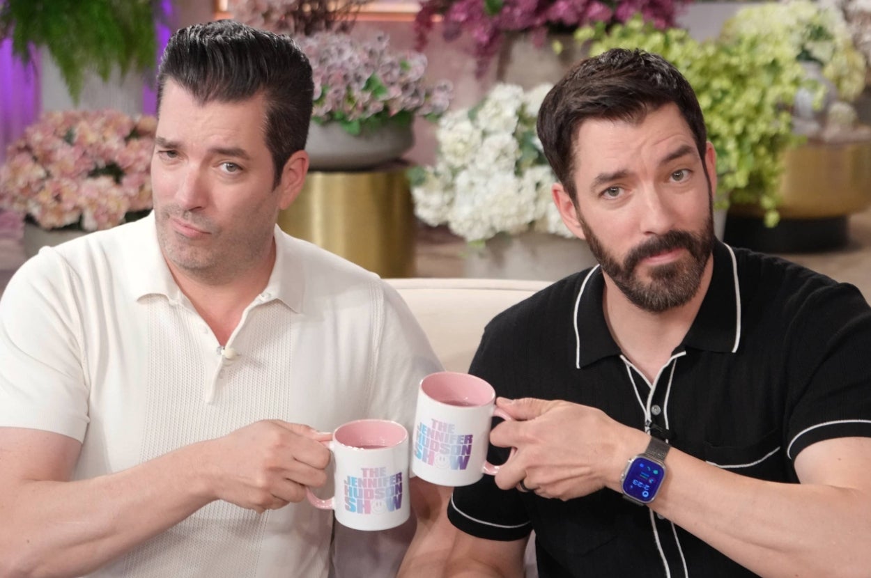 Two men with beards, casually dressed, holding matching mugs on a talk show set with floral decor in the background