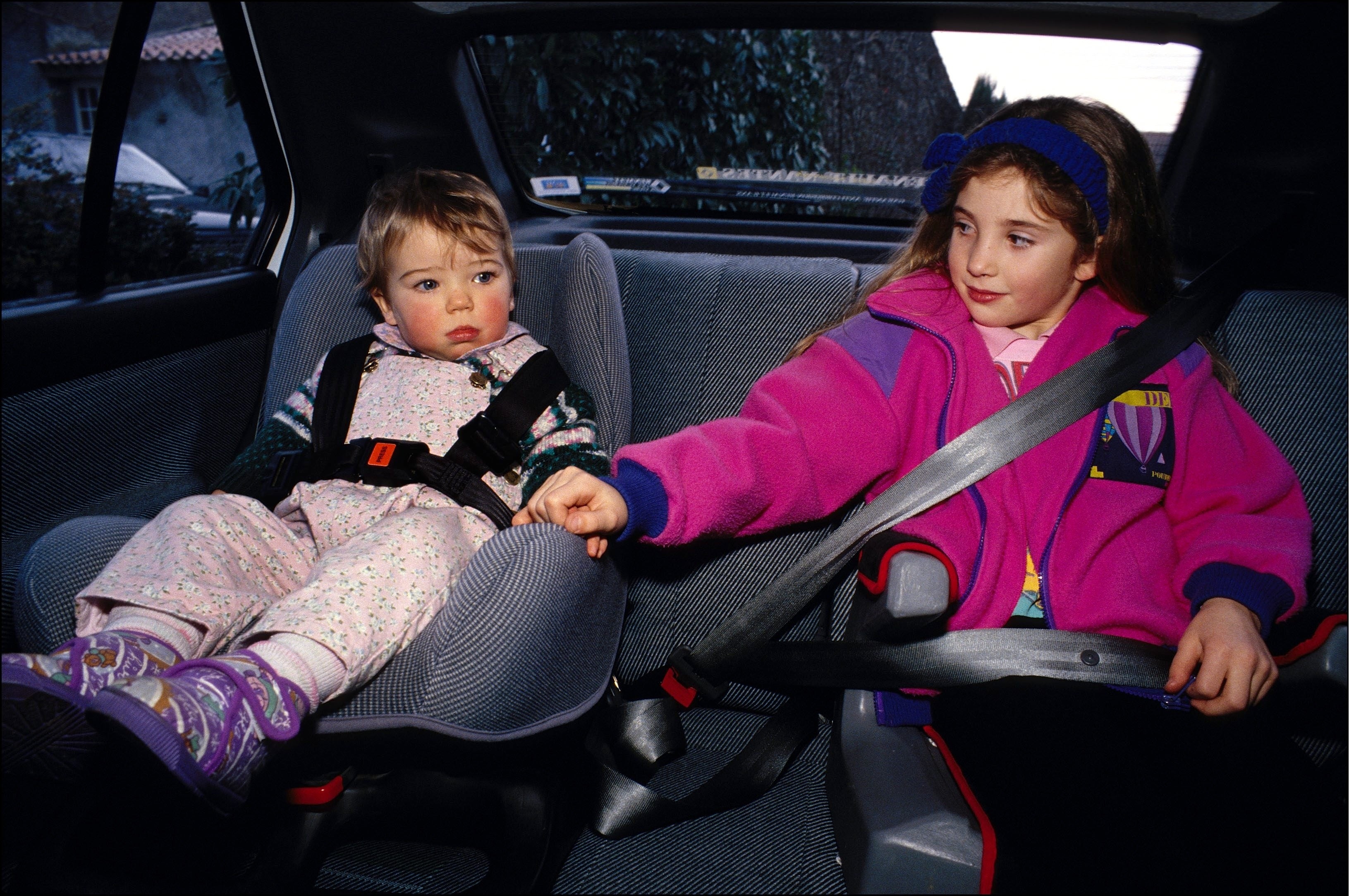Two children in the backseat of a car, one in a car seat and the other buckled in, holding hands