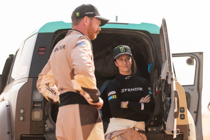 Two people in racing gear, one leaning against a vehicle, appear relaxed and in conversation by the open back of an SUV or racing vehicle