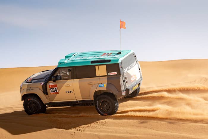 Off-road vehicle driving through desert dunes, kicking up sand. It has a roof flag and sponsor logos on the side