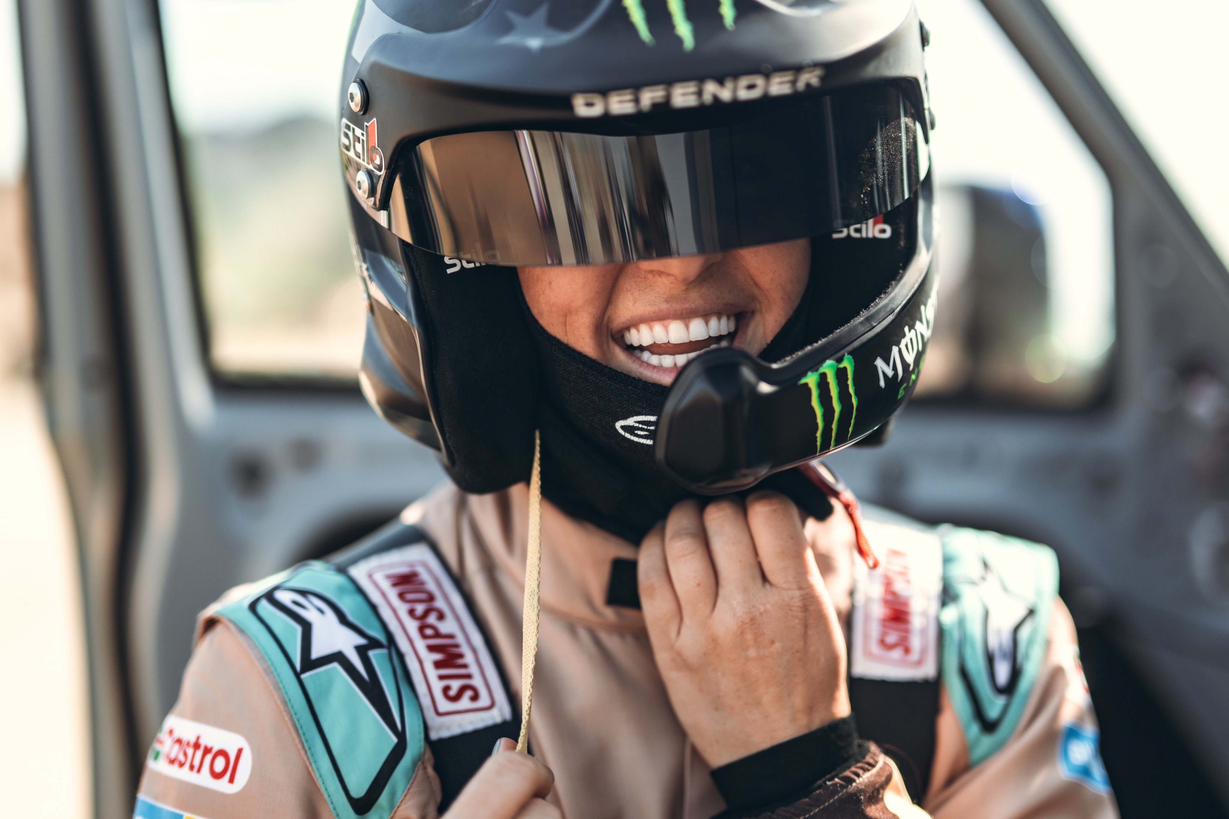 Person smiling while adjusting a racing helmet, wearing a suit with sponsor patches