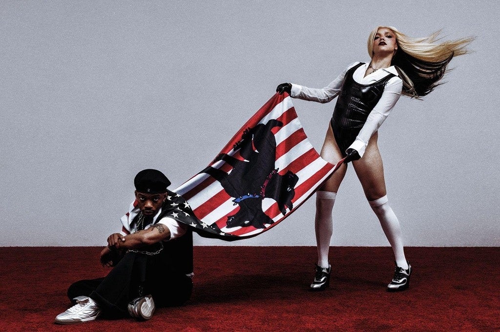 Two performers pose dramatically; one stands in a bodysuit and boots holding an American flag with a silhouette, while the other sits casually on the floor