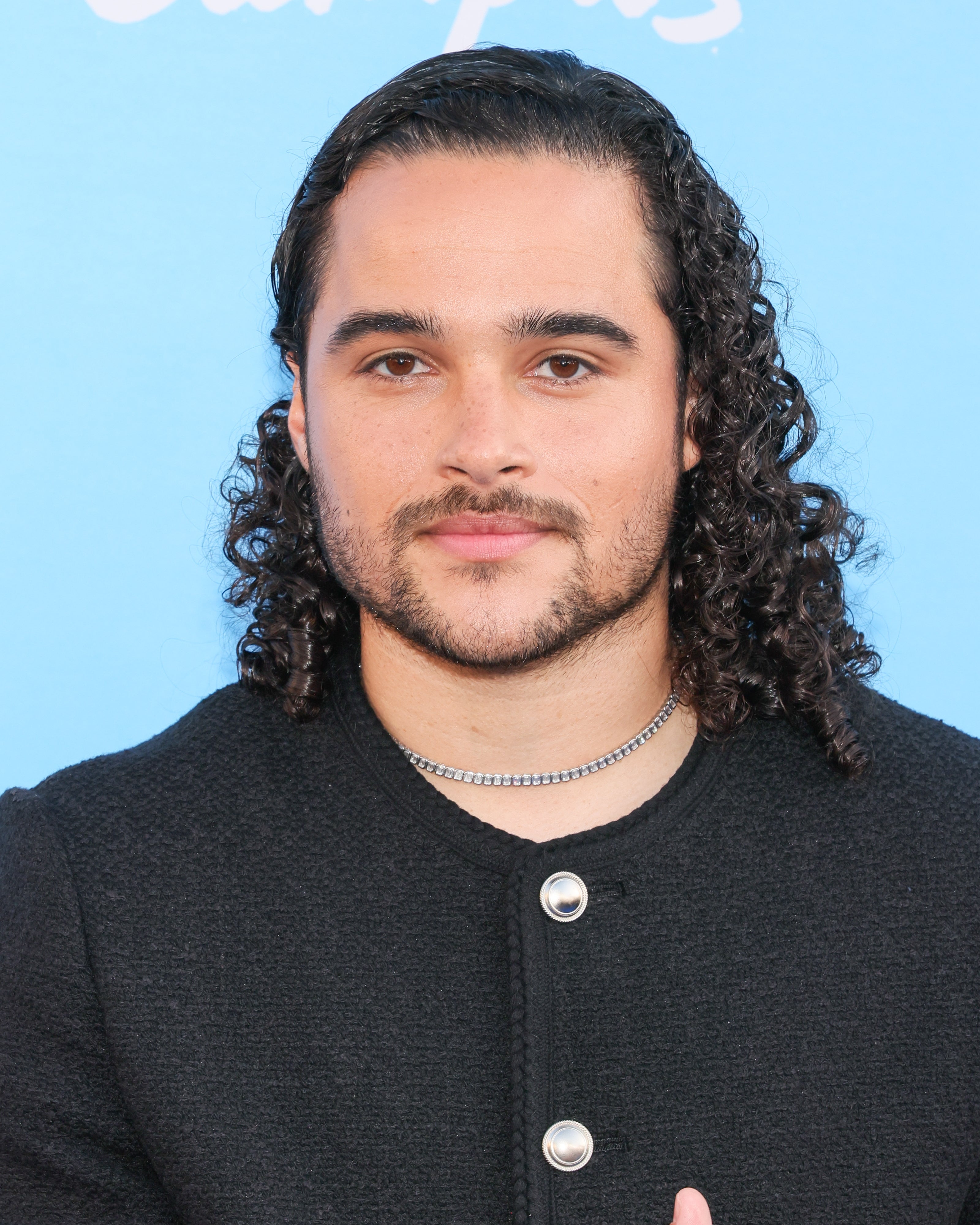 A person with curly hair wears a black buttoned jacket and a silver necklace, posing against a plain backdrop