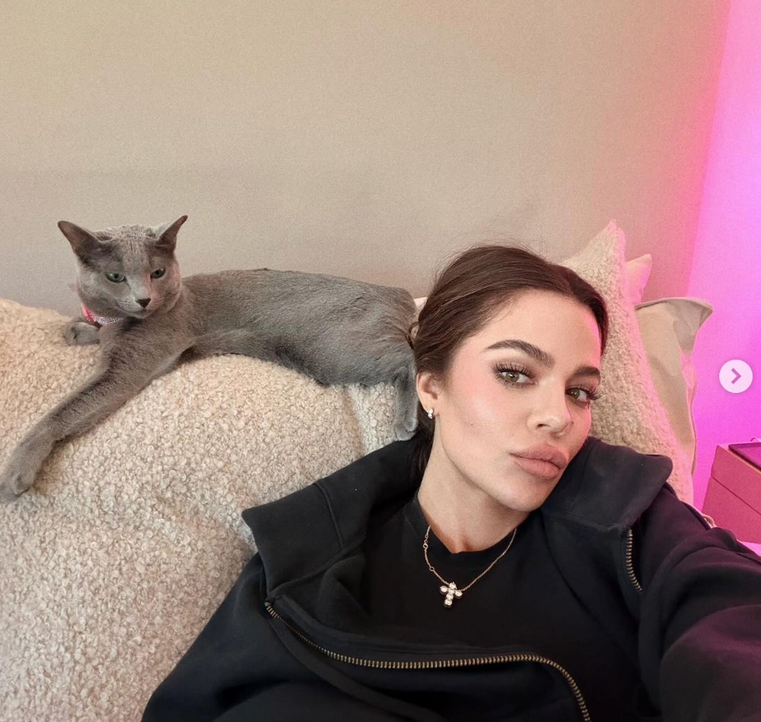 Person lounging with a sleek cat, wearing casual clothing and a necklace, posing indoors