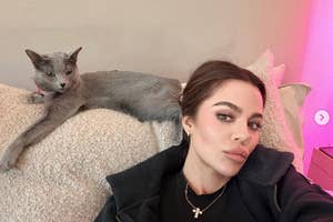 Person lounging with a sleek cat, wearing casual clothing and a necklace, posing indoors