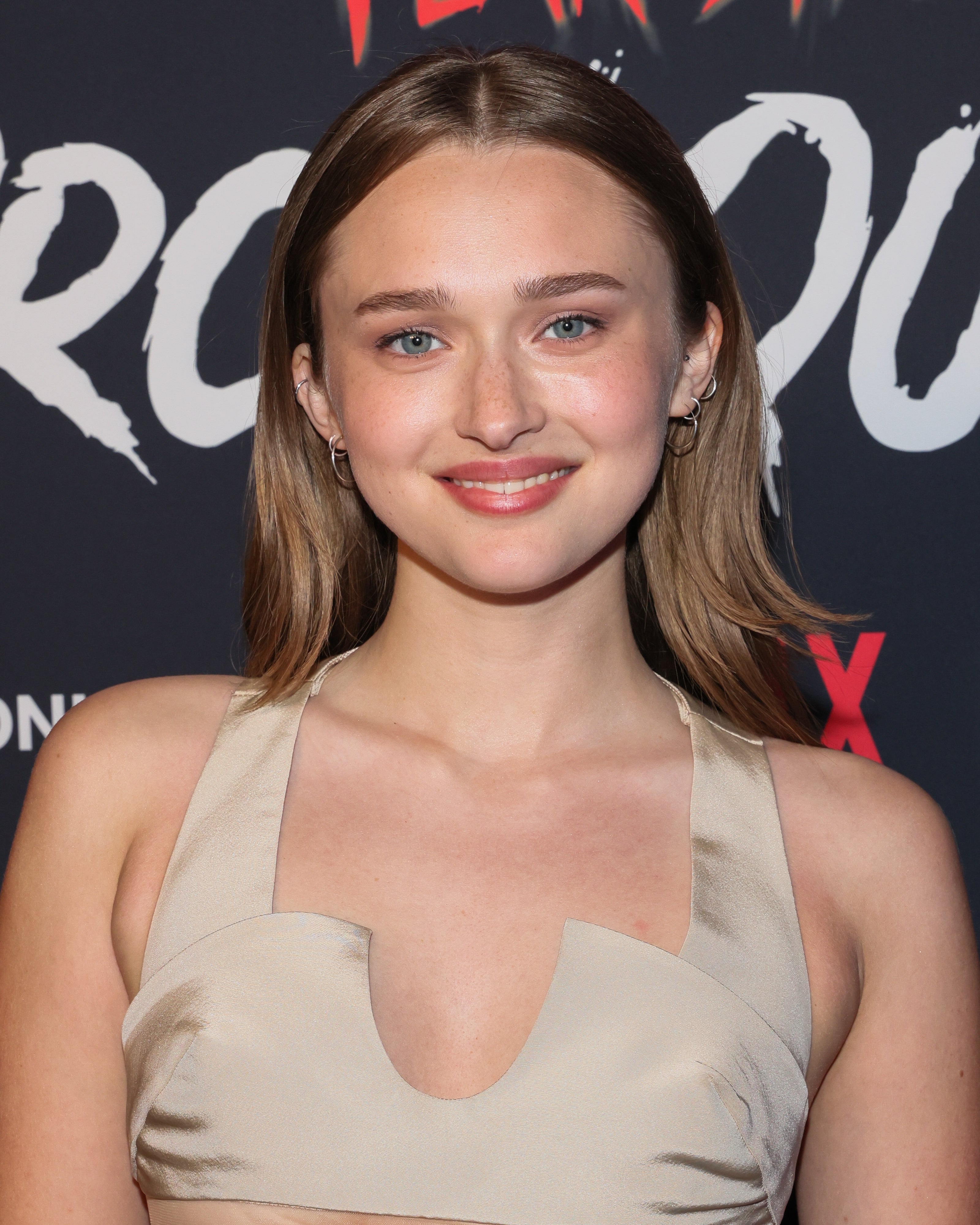 Person smiling at a TV event, wearing a sleeveless, unique-cut dress with a fitted bodice, standing in front of a Netflix-themed backdrop