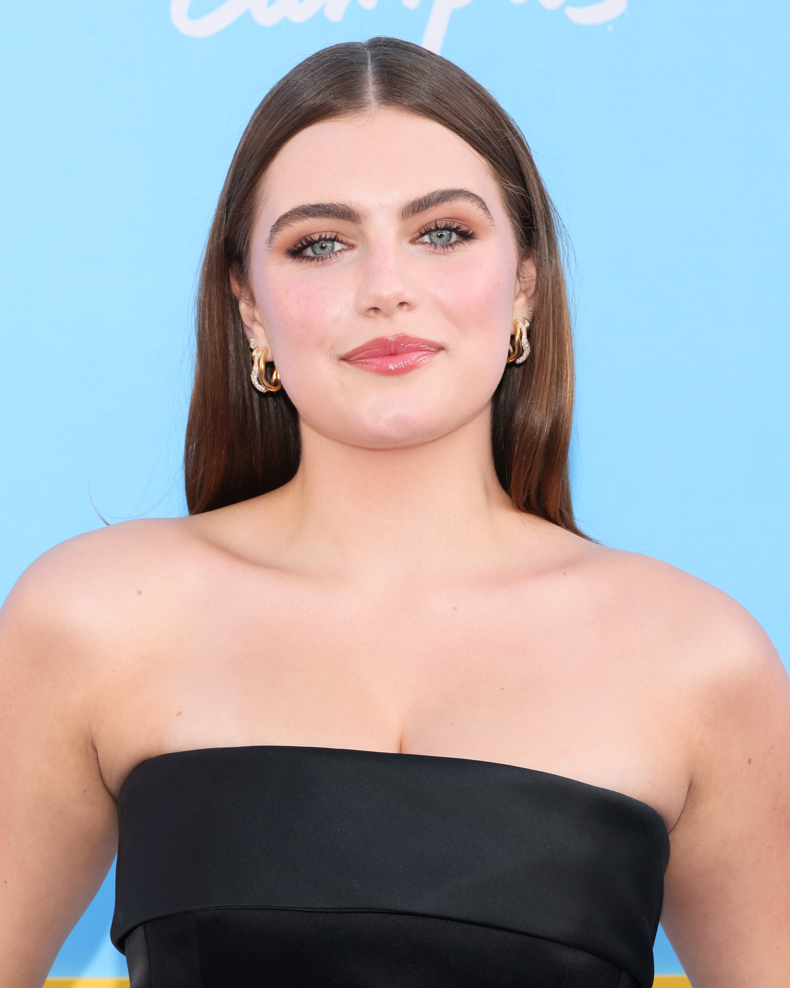 Person smiling on a red carpet, wearing a sleek strapless outfit and gold earrings