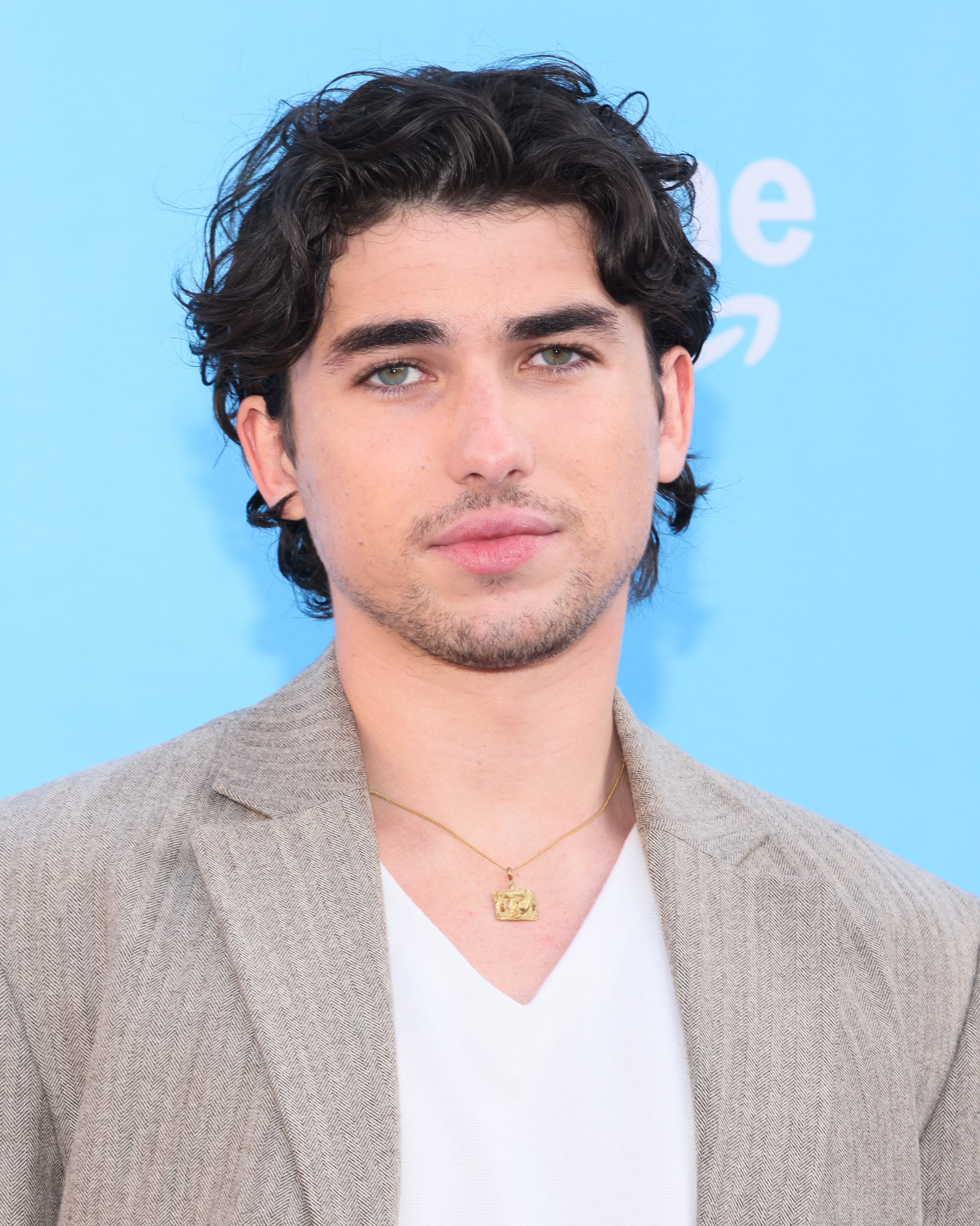 Person wearing a beige blazer and white top, accessorized with a gold necklace, posing in front of a light blue background