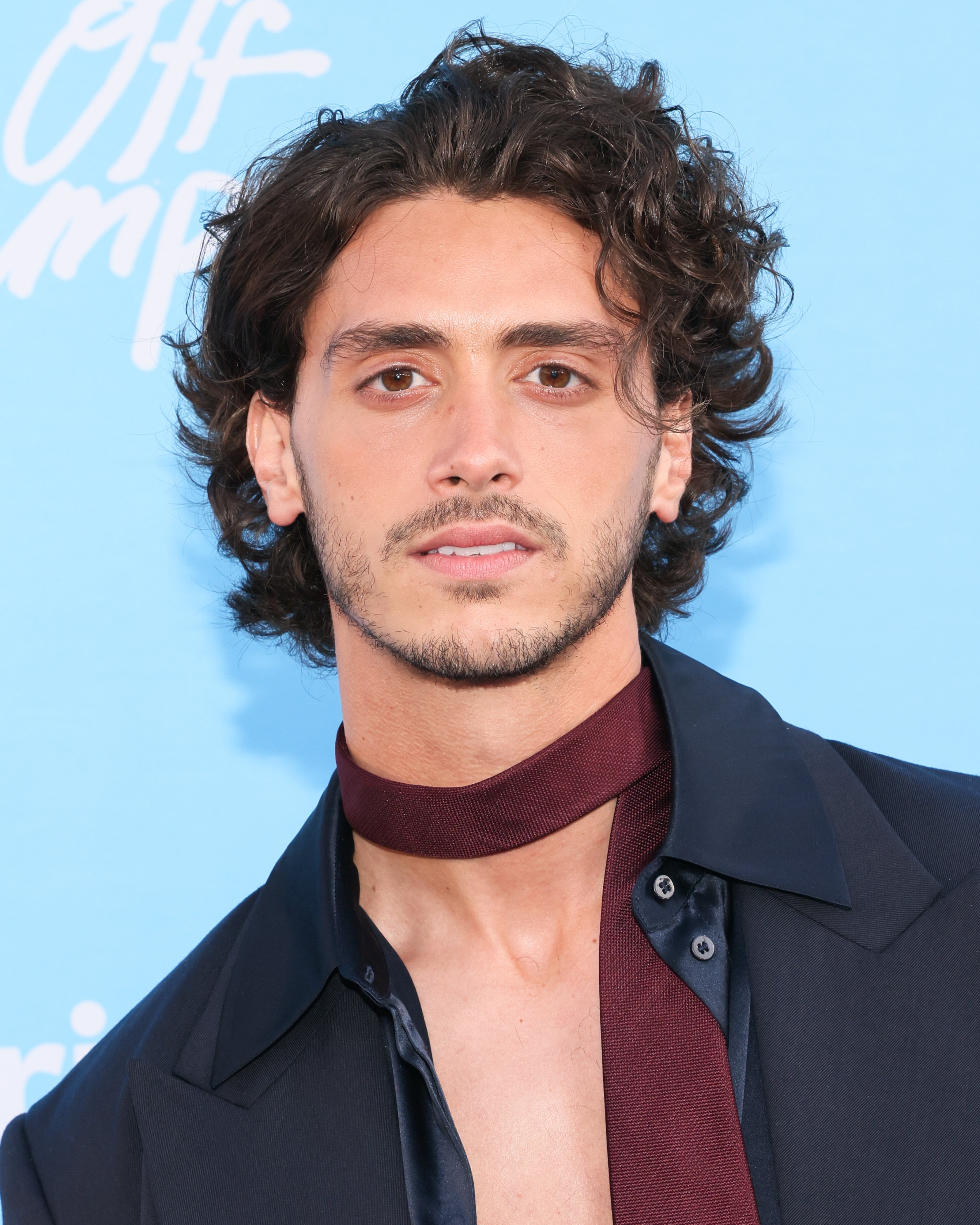 Person on red carpet wearing a dark open shirt with a burgundy scarf. Background has text with unclear details