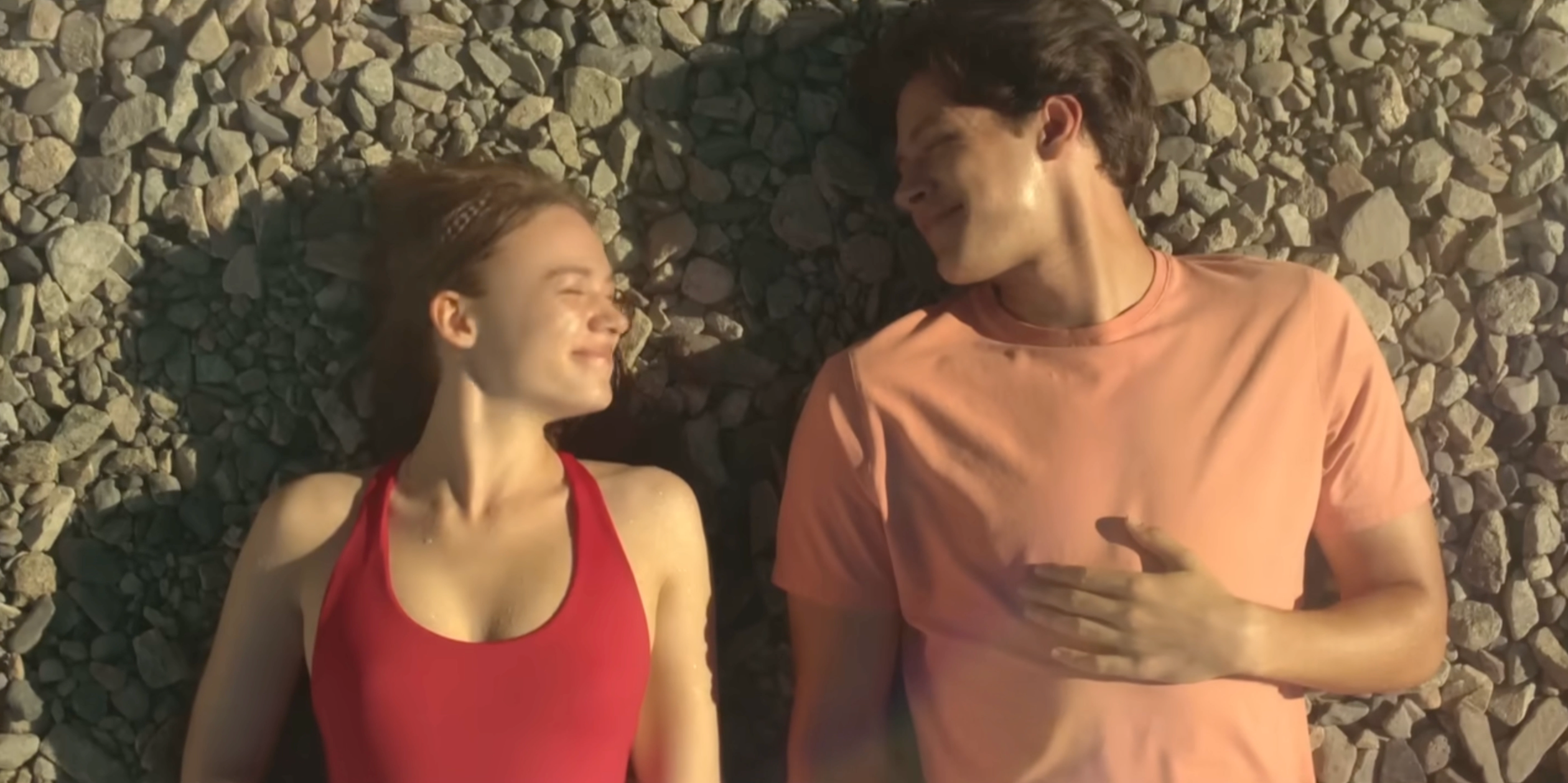 Two people lay on a pebbled surface, facing each other, smiling. The person on the left wears a sleeveless top, while the person on the right wears a T-shirt