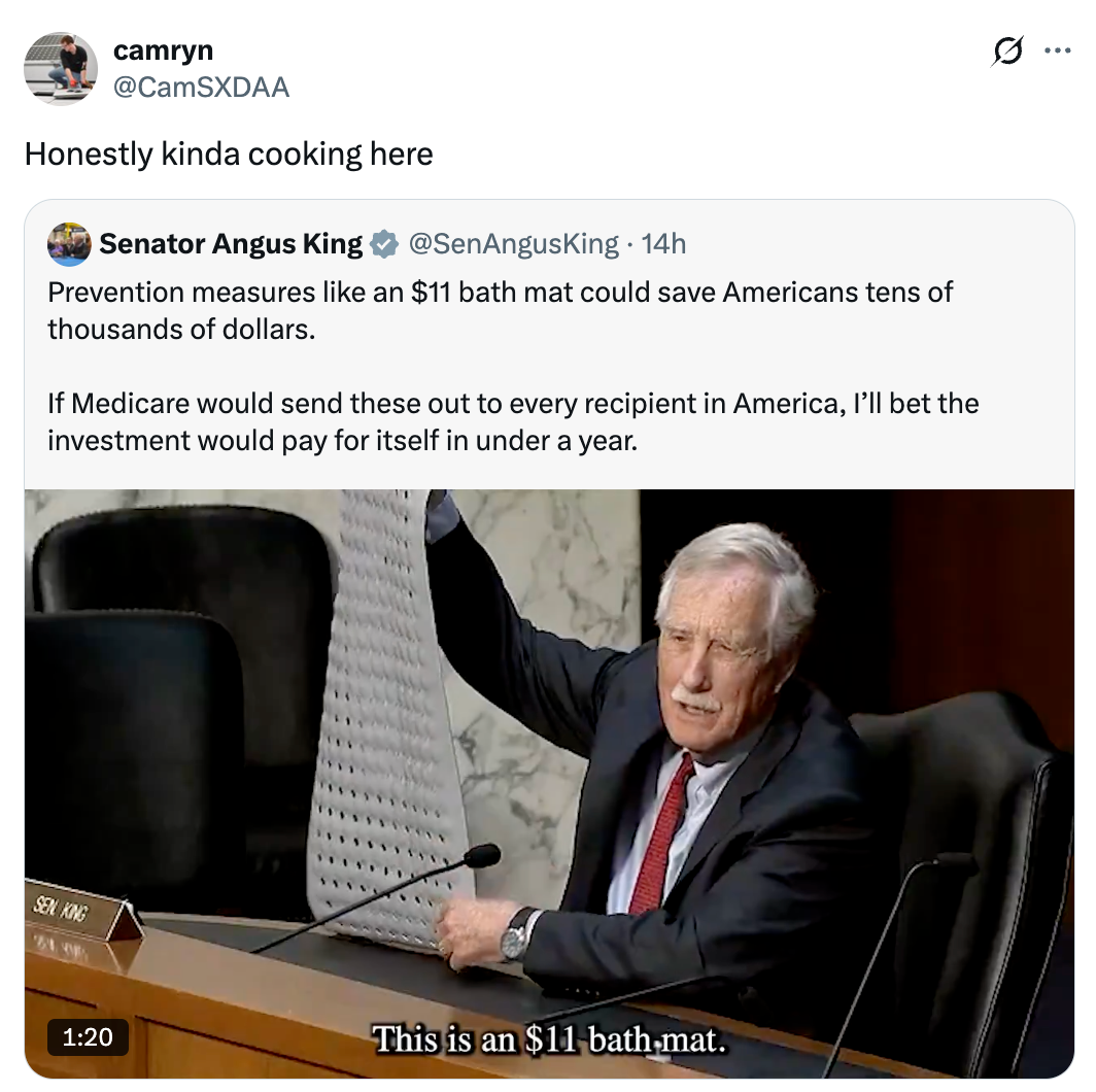 Man in a suit holding up a bath mat in a hearing, discussing cost-saving measures with printed text overlay