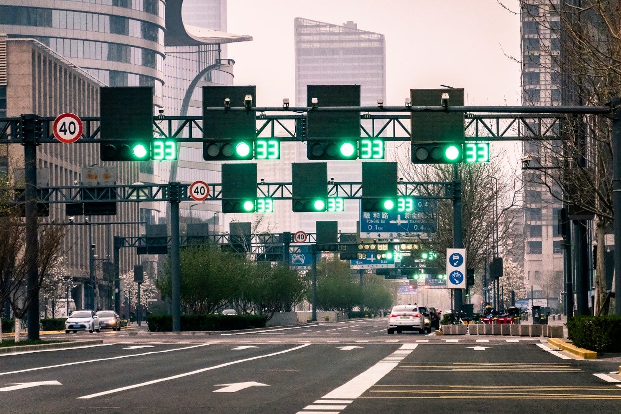 City street with green traffic lights, few cars, and modern buildings. Sparse traffic and an overpass are visible in the urban setting