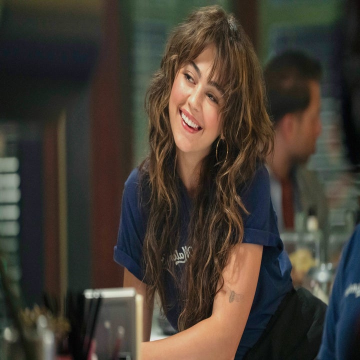 Pessoa sorrindo casualmente dentro de casa, vestindo uma camiseta azul, com cabelos longos e ondulados. Em um ambiente social com figuras desfocadas ao fundo
