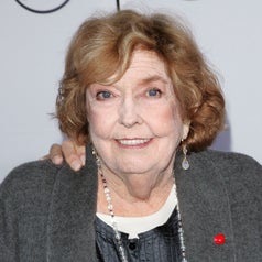Anne Meara smiling at an event, wearing an outfit with a beaded necklace and a small strawberry shaped pin