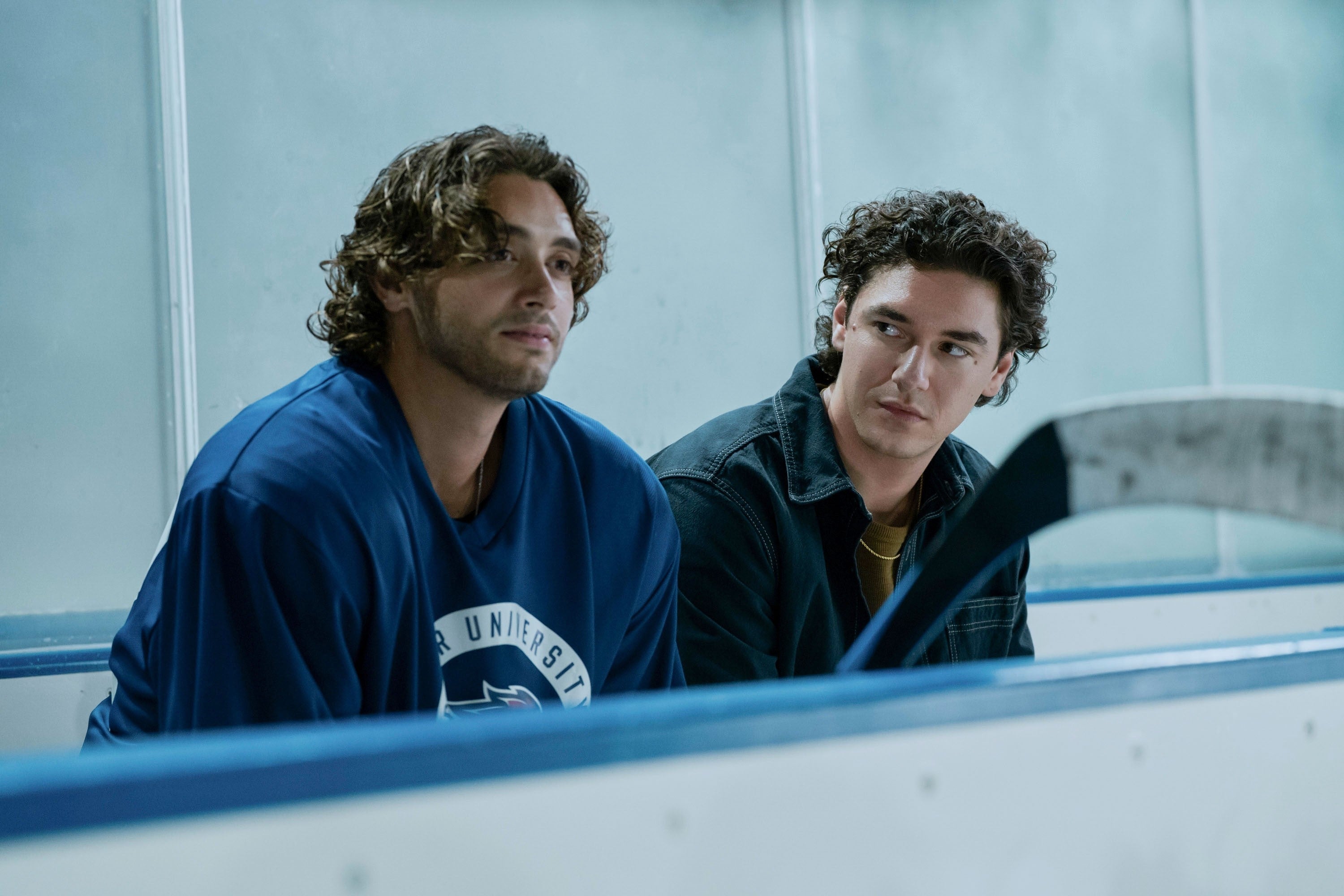 Two men sit on a hockey bench, one in a jersey and the other in casual attire, engaged in a serious conversation