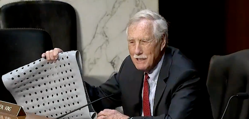 Person holding up a large grid sheet during a formal meeting, seated at a table with microphones and nameplate