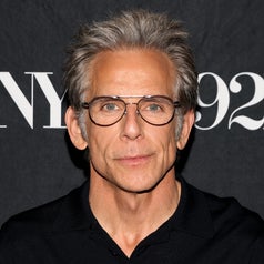 Ben Stiller in a shirt with glasses poses in front of a backdrop with text