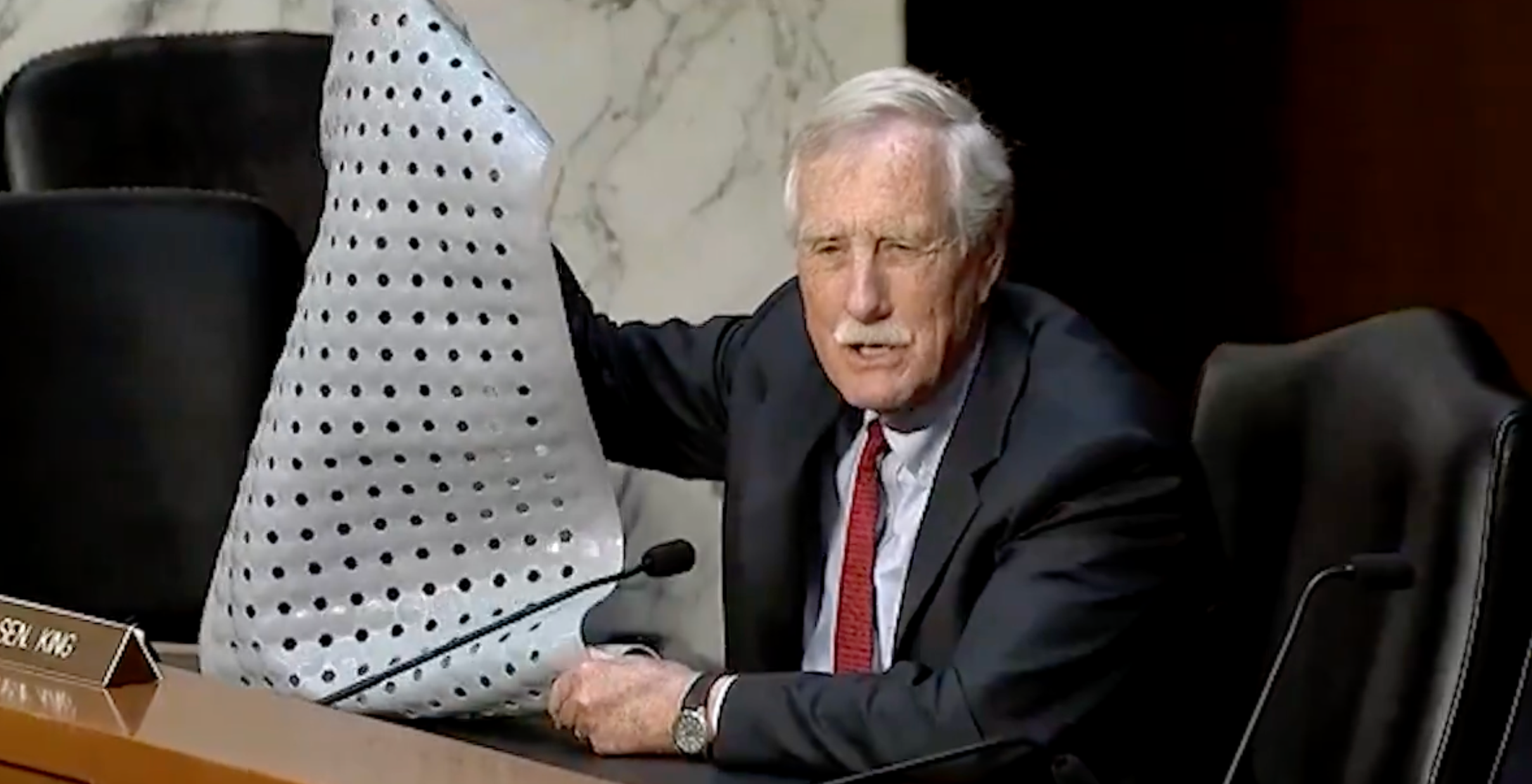 Person in a suit holds up a sheet with holes during a hearing or meeting, speaking into a microphone