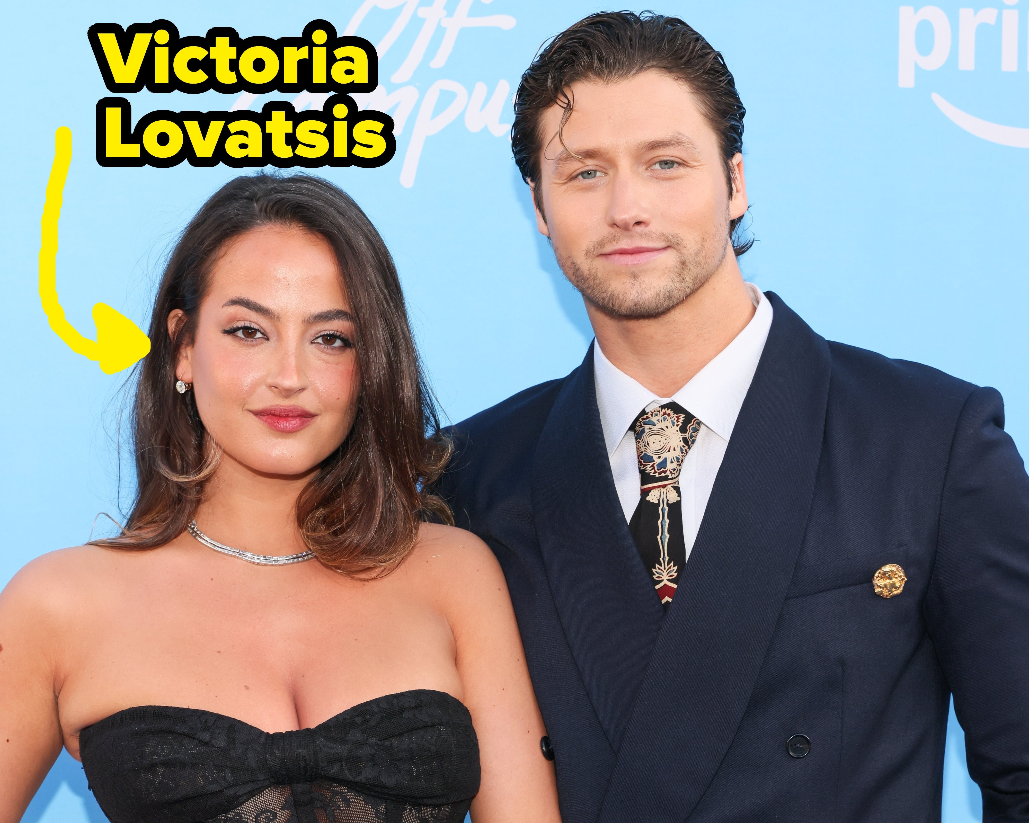 Two people on the red carpet; the woman wears a strapless lace dress, and the man dons a formal suit with a patterned tie