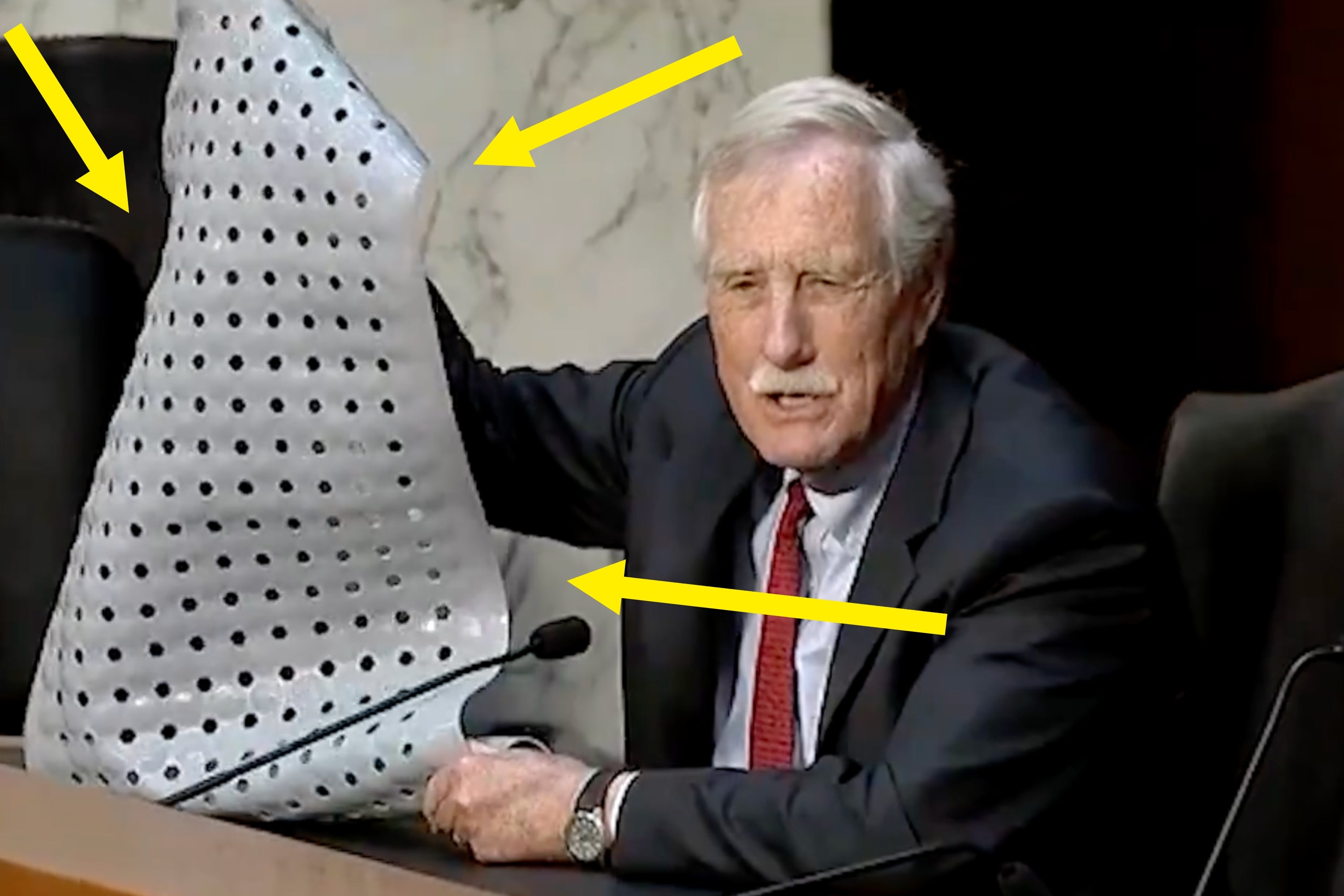 Person in a suit holds up a sheet with holes during a hearing or meeting, speaking into a microphone