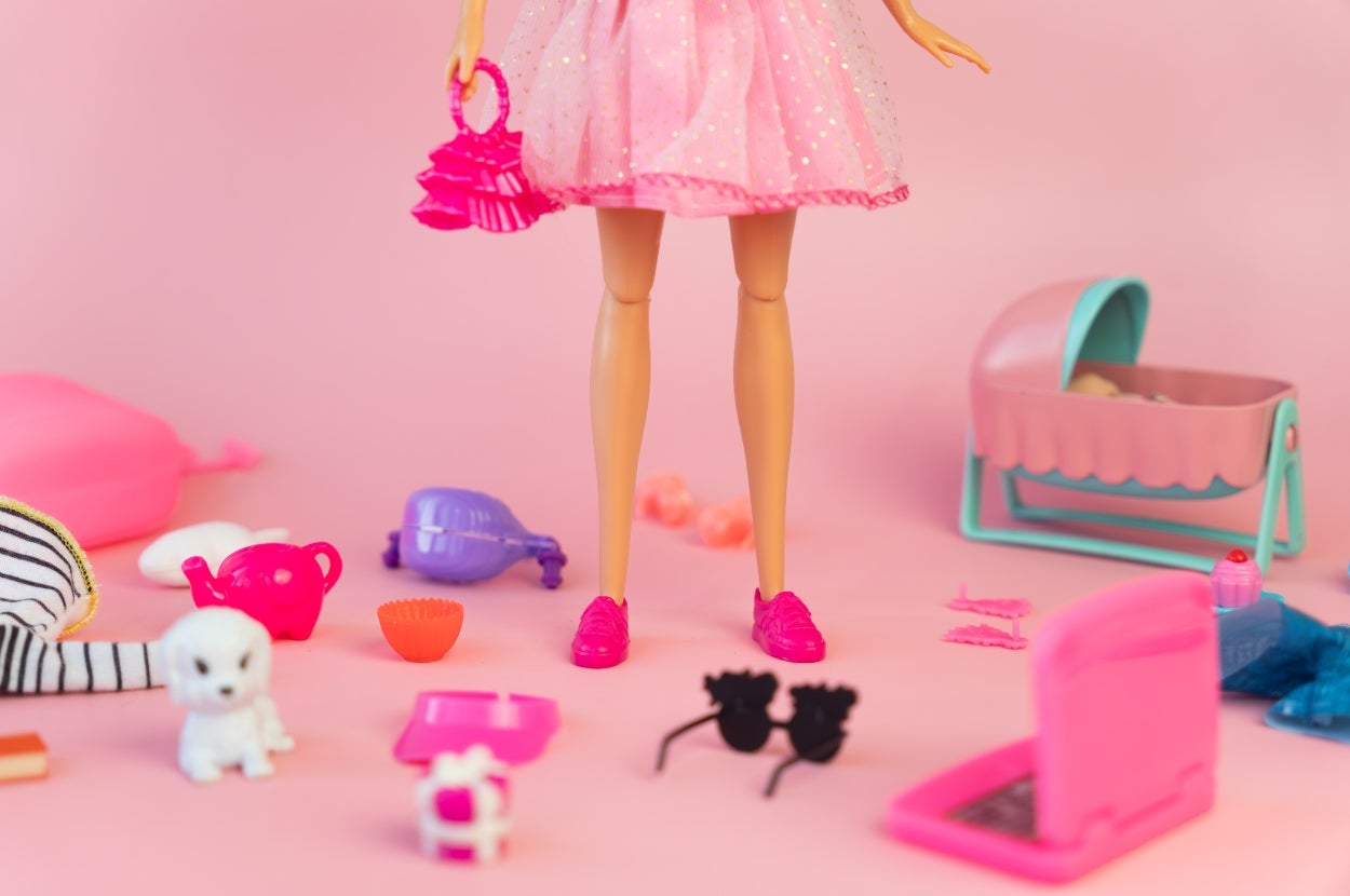 A doll in a pink dress holds a pink handbag, surrounded by small toys and accessories, including a laptop, sunglasses, and a puppy figure