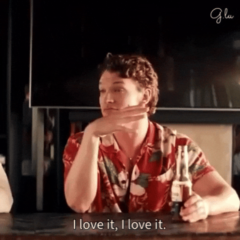 Person in a floral shirt at a bar, smiling and gesturing with a beer, saying "I love it, I love it."