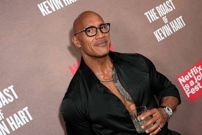 Man with glasses in an open-collar shirt holds a drink at "The Roast of Kevin Hart" event backdrop