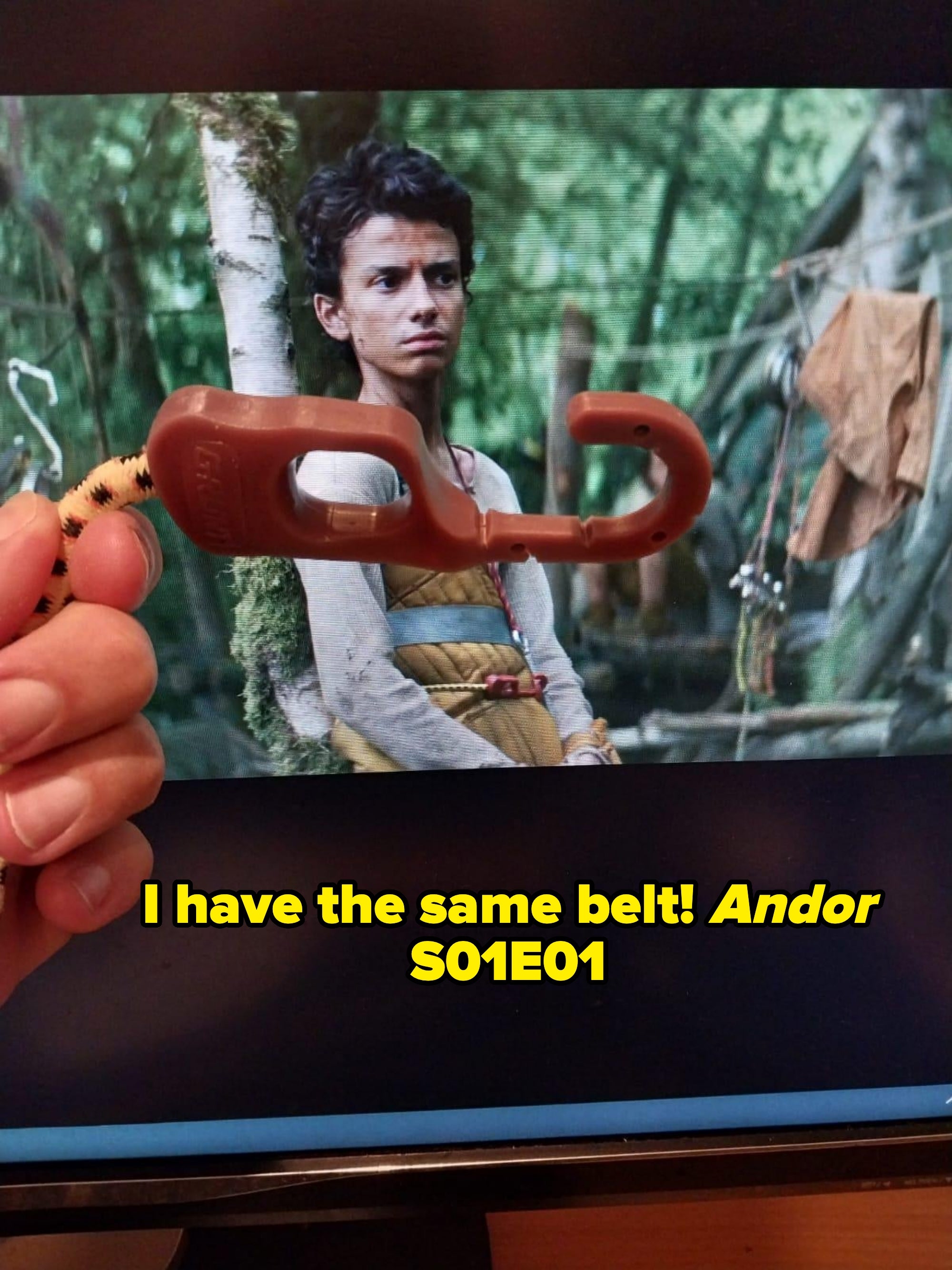 A hand holds a toy hook in front of a scene from a medieval-style TV show or movie, featuring a person in rustic attire in a forest setting