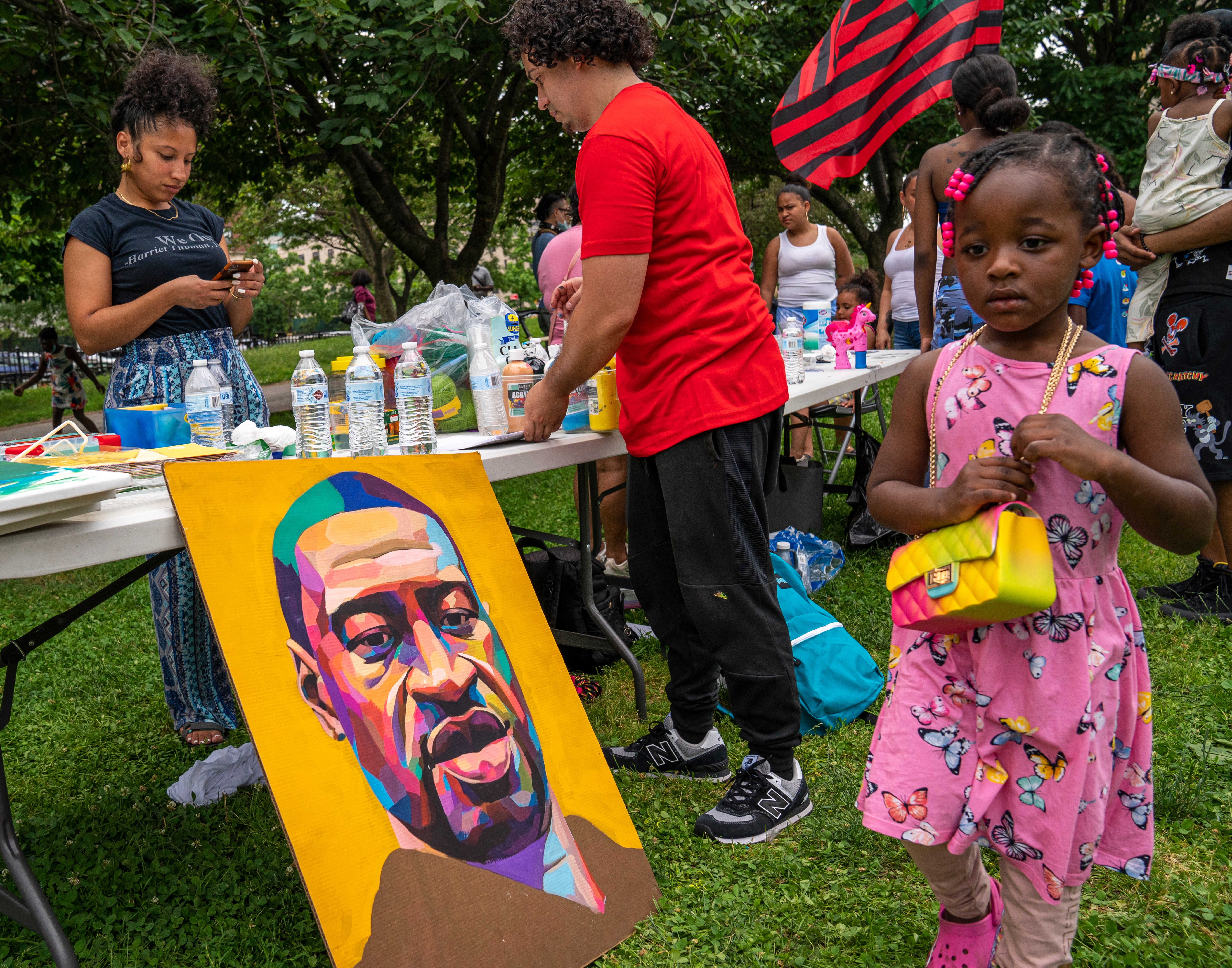 A kid successful a pinkish formal holds a artifact portion radical stitchery nearby. A colorful representation of George Floyd is propped up connected the grass