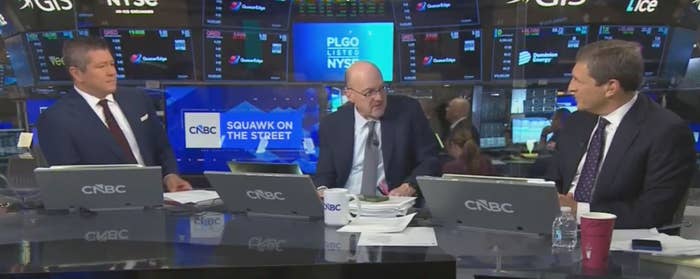 Three news anchors are seated at a CNBC desk discussing financial news in a stock market trading room. Monitors display various stock data