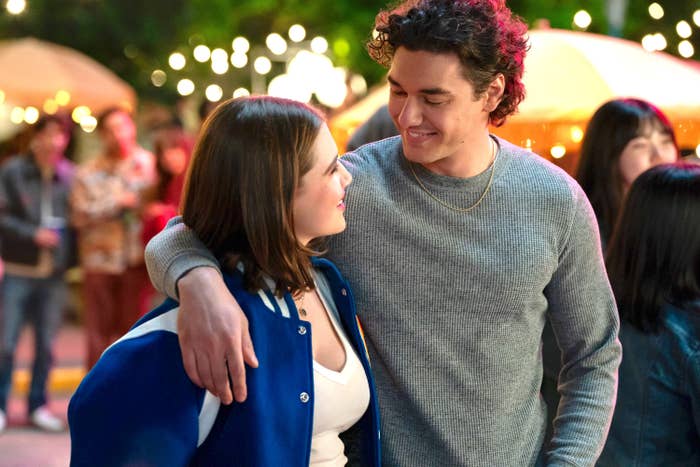 Two actors smiling at each other, with one in a varsity jacket and the other in a casual sweater, on a lively outdoor set