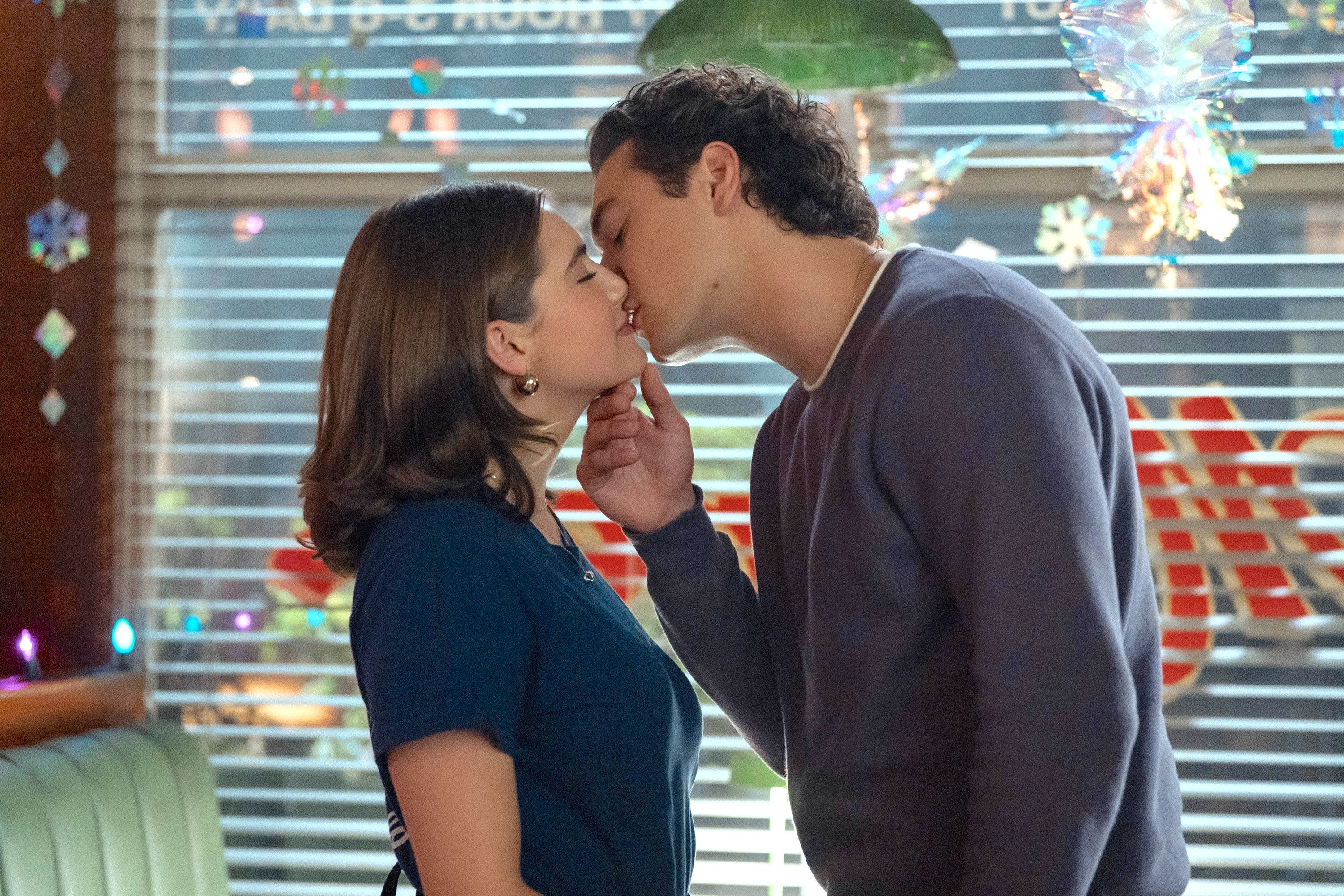 Two people kiss in a diner setting, both casually dressed. Holiday decorations are visible in the background