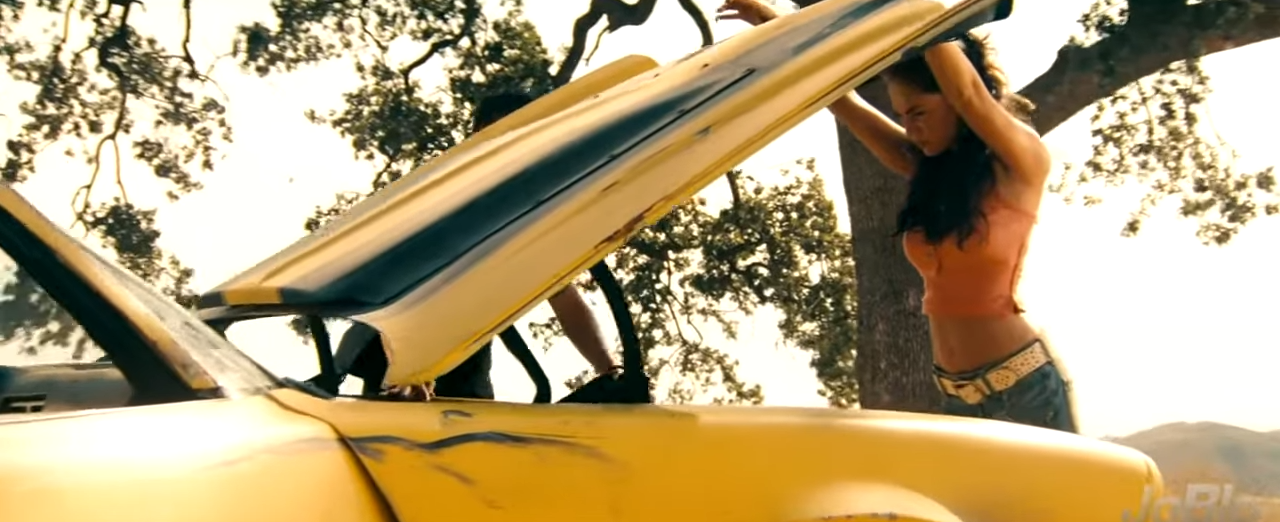 Person standing by a yellow car with an open hood, under a tree, adjusting something on their head