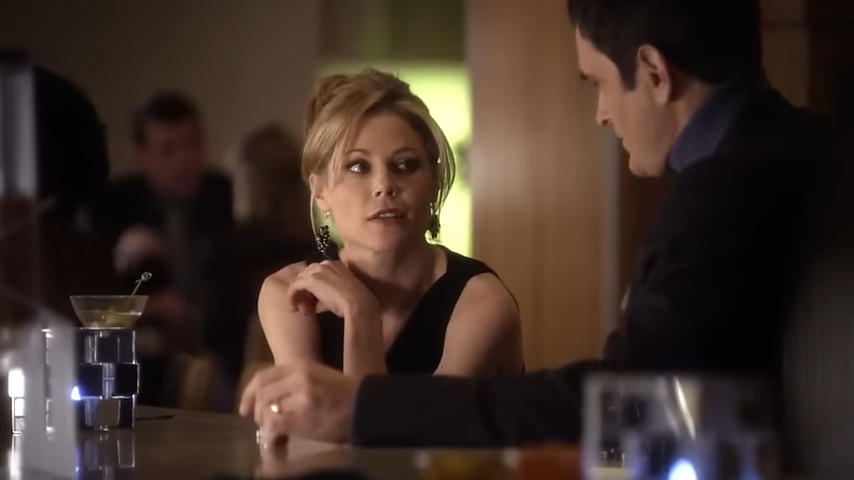 A woman in elegant attire sits at a bar, engaged in conversation with a man in a suit