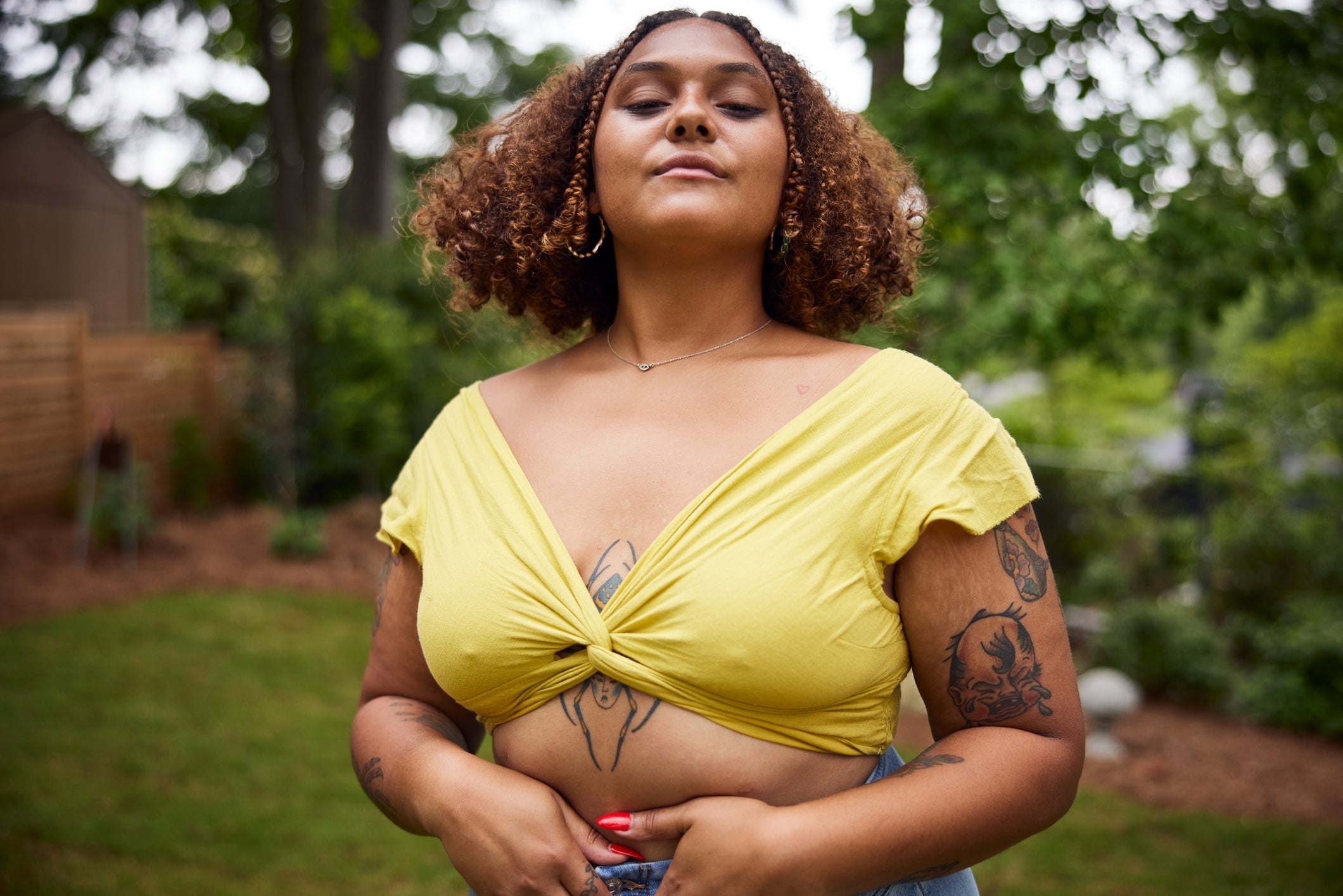 Person confidently poses outdoors in a twisted top with floral tattoos visible on arms and chest