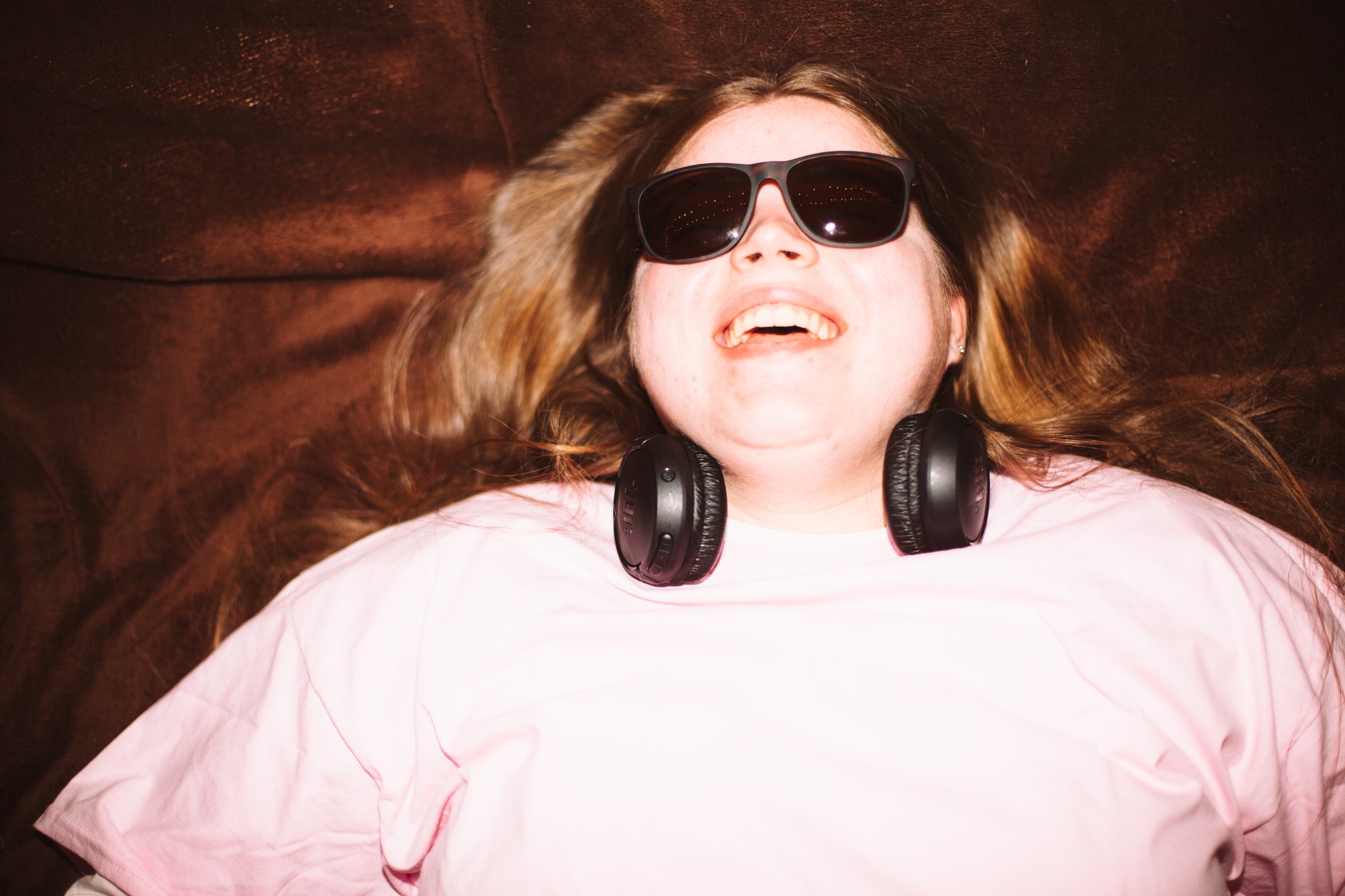 Person lying down, smiling, wearing sunglasses and headphones around their neck, conveying relaxation and enjoyment