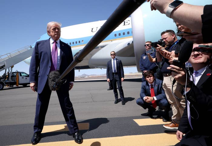 Person in suit speaks to media on airport tarmac with airplane in background. Journalists hold microphones and recorders