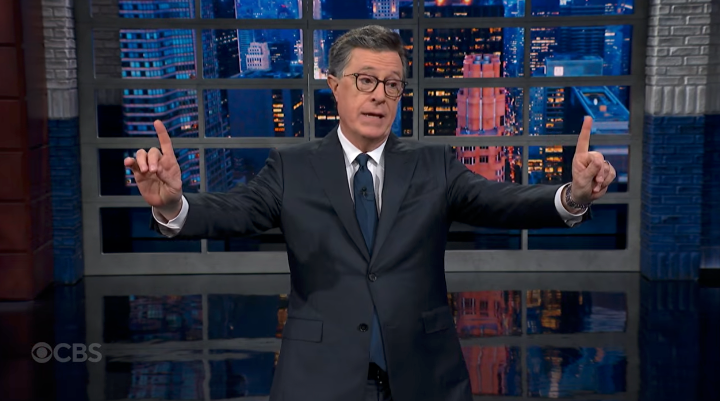 A man in a suit gestures with both index fingers up, standing on a talk show set with a cityscape background