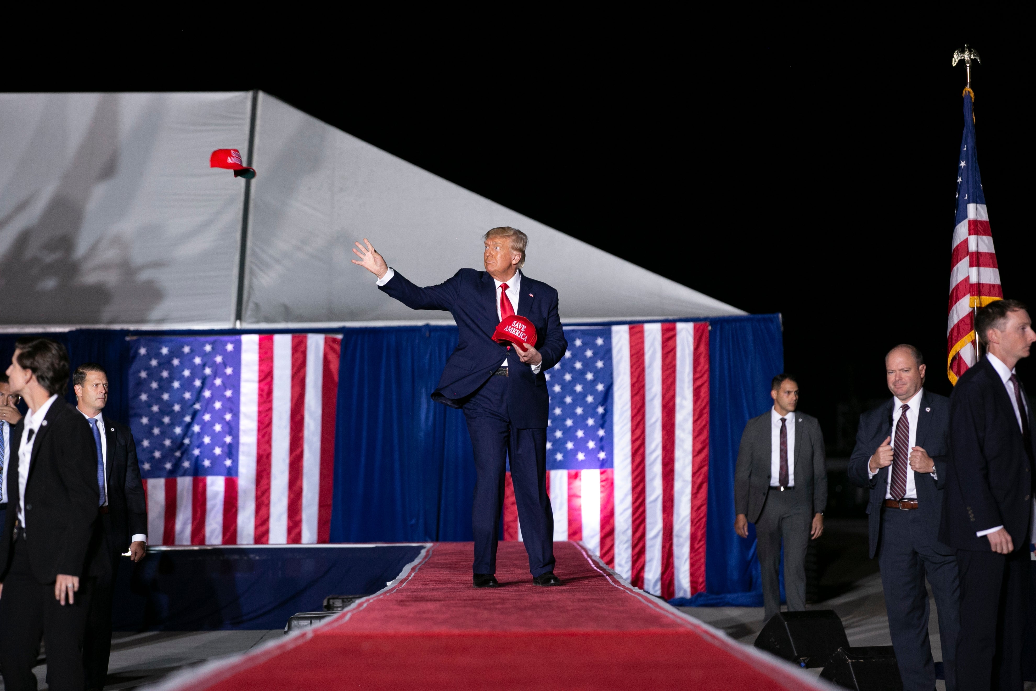 A man in a suit throws a hat on stage at an event, surrounded by people in suits, with American flags in the background