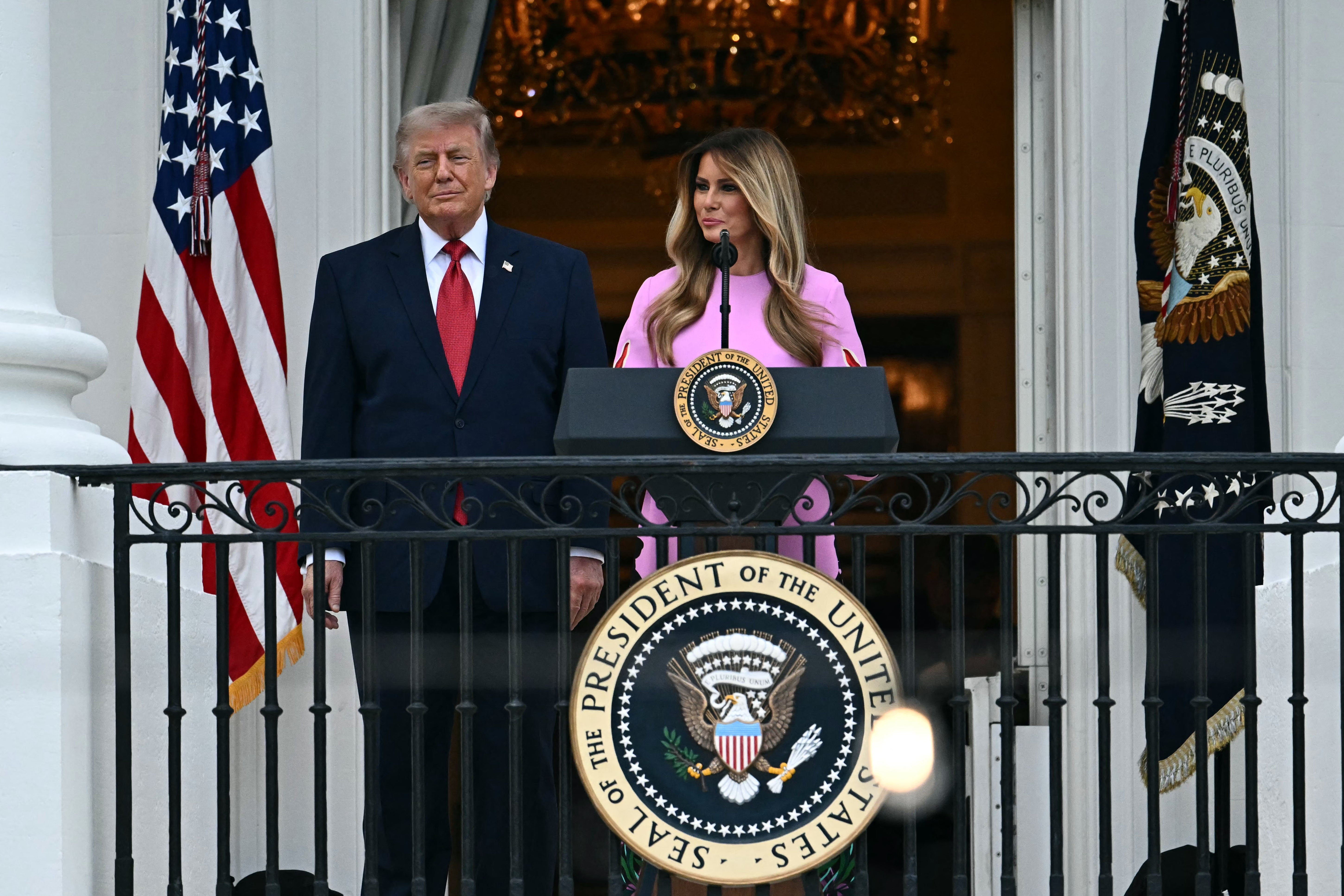 Two individuals stand on a balcony with the U.S. presidential seal, speaking at a podium, with American flags in the background