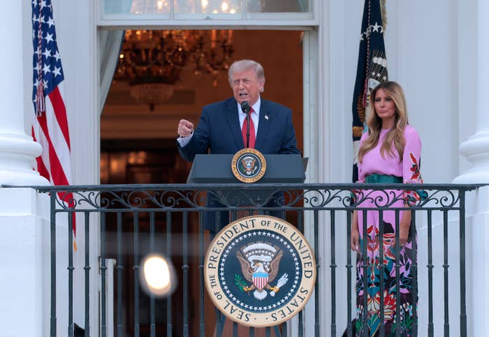 A antheral   speaks astatine  a podium connected  a balcony with a seal of the President of the United States. A pistillate   stands beside him wearing a floral dress