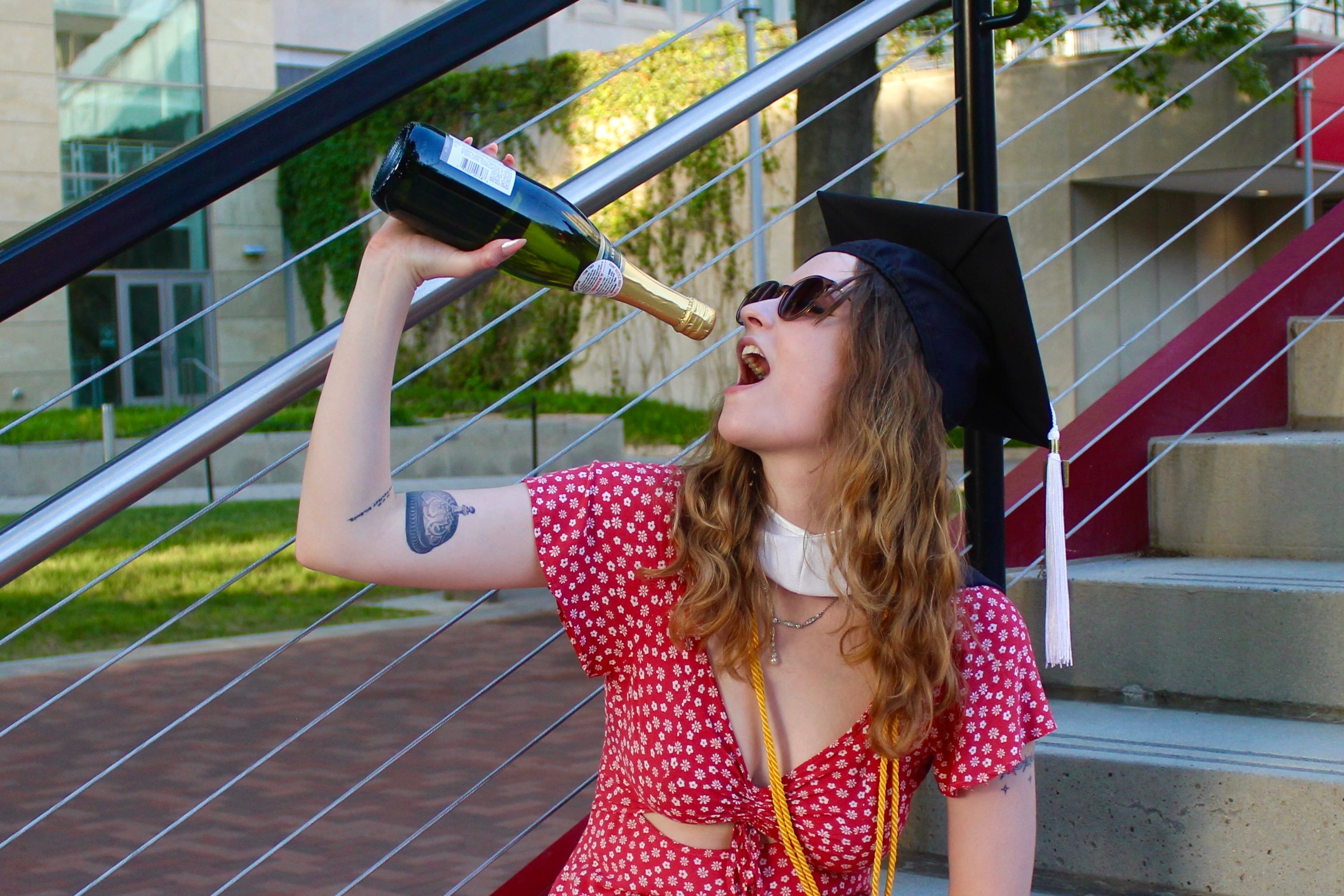 Me wearing a grad cap and sunglasses while pretending to drink from a bottle of champagne on an outdoor staircase